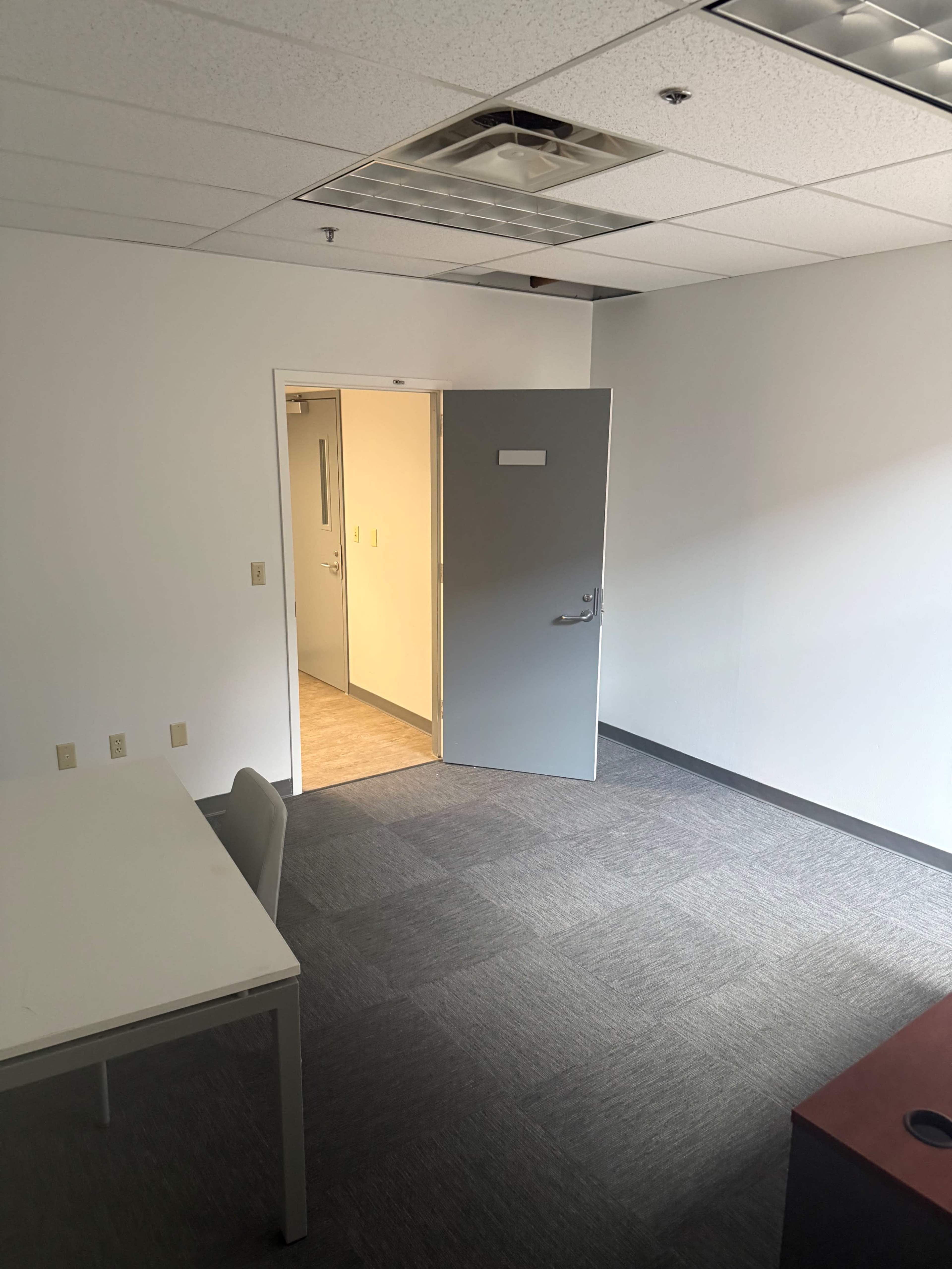 The image shows a sparsely furnished office with a table and chair, a doorway leading to another room, and gray carpet flooring.