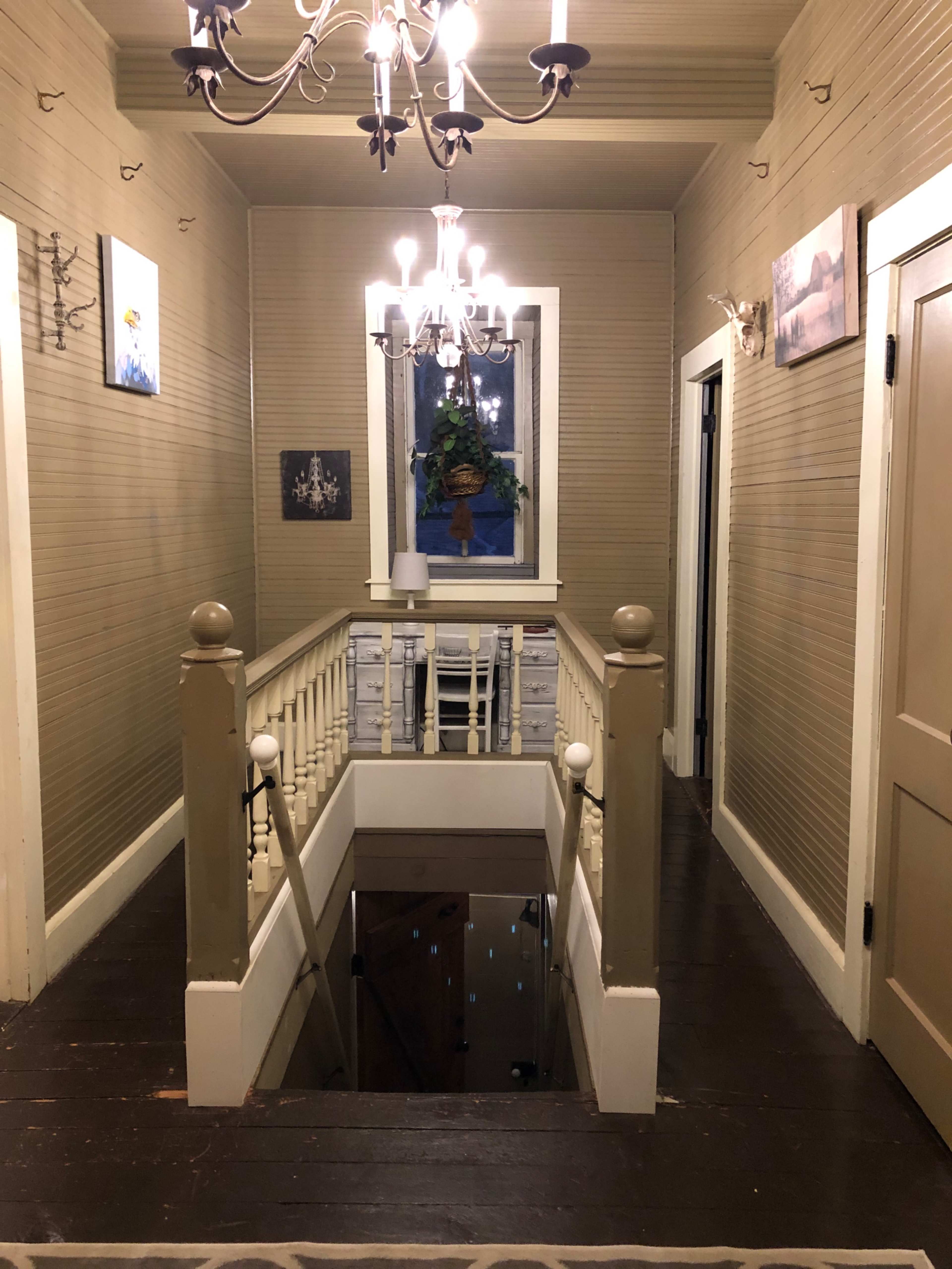 The image shows a hallway with a chandelier, a staircase railing, and framed pictures on the walls, leading to a window with a potted plant.