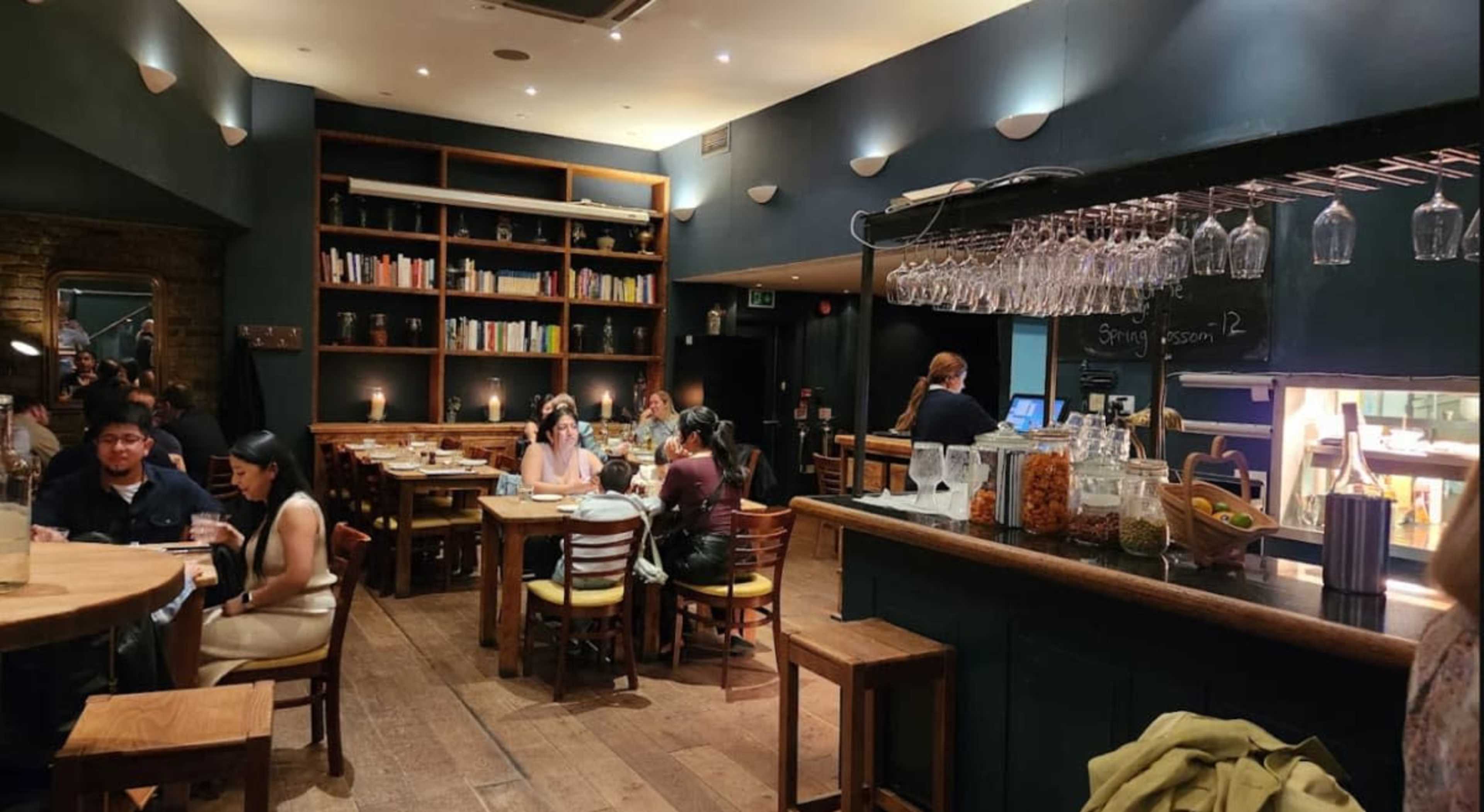 The image shows a crowded restaurant interior with wooden tables and shelves lined with books, where diners are seated and a counter displays glassware and jars.