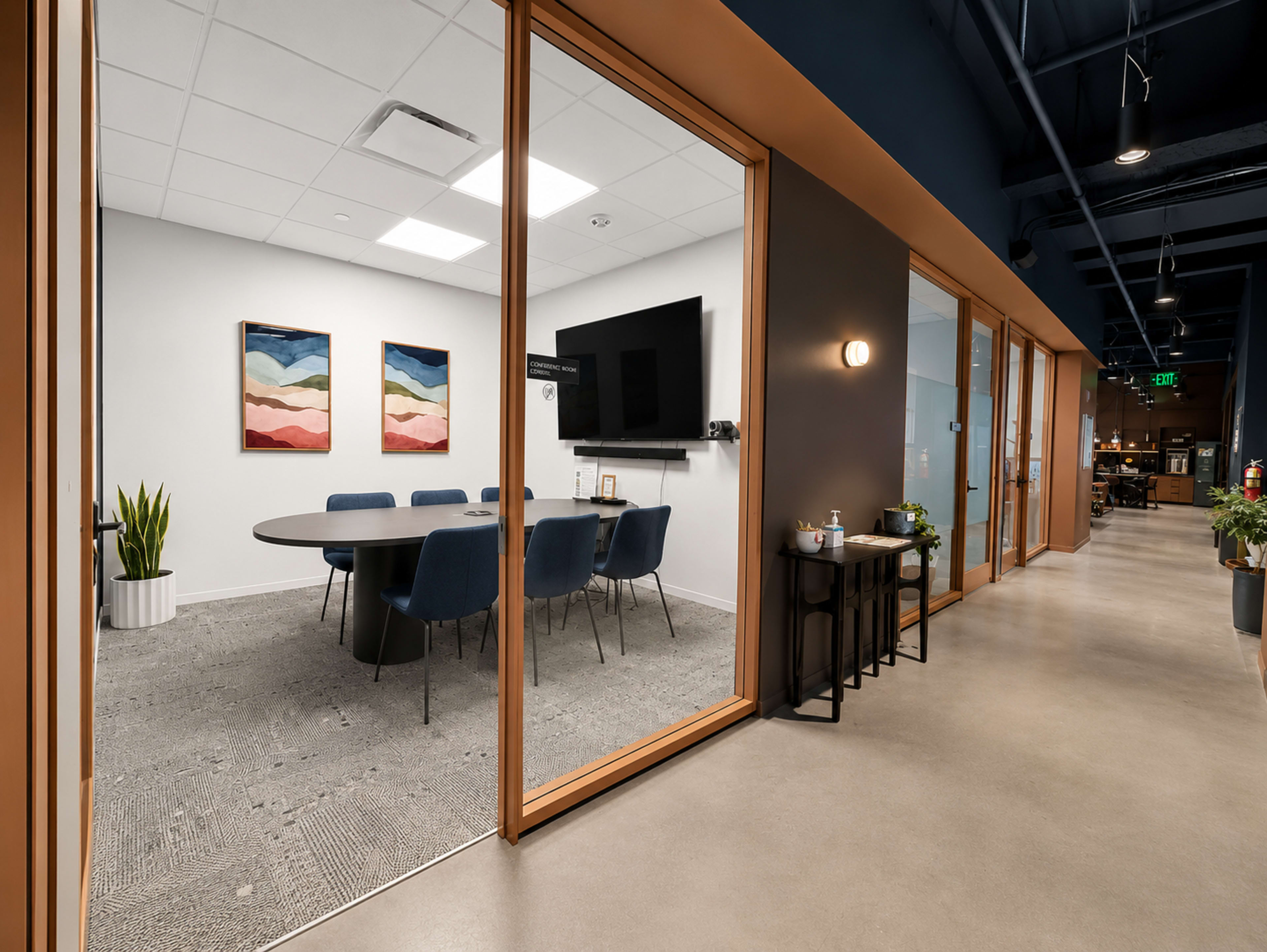 The image shows a modern office corridor leading to a glass-enclosed meeting room with a round table and upholstered chairs, featuring colorful artwork on the walls.
