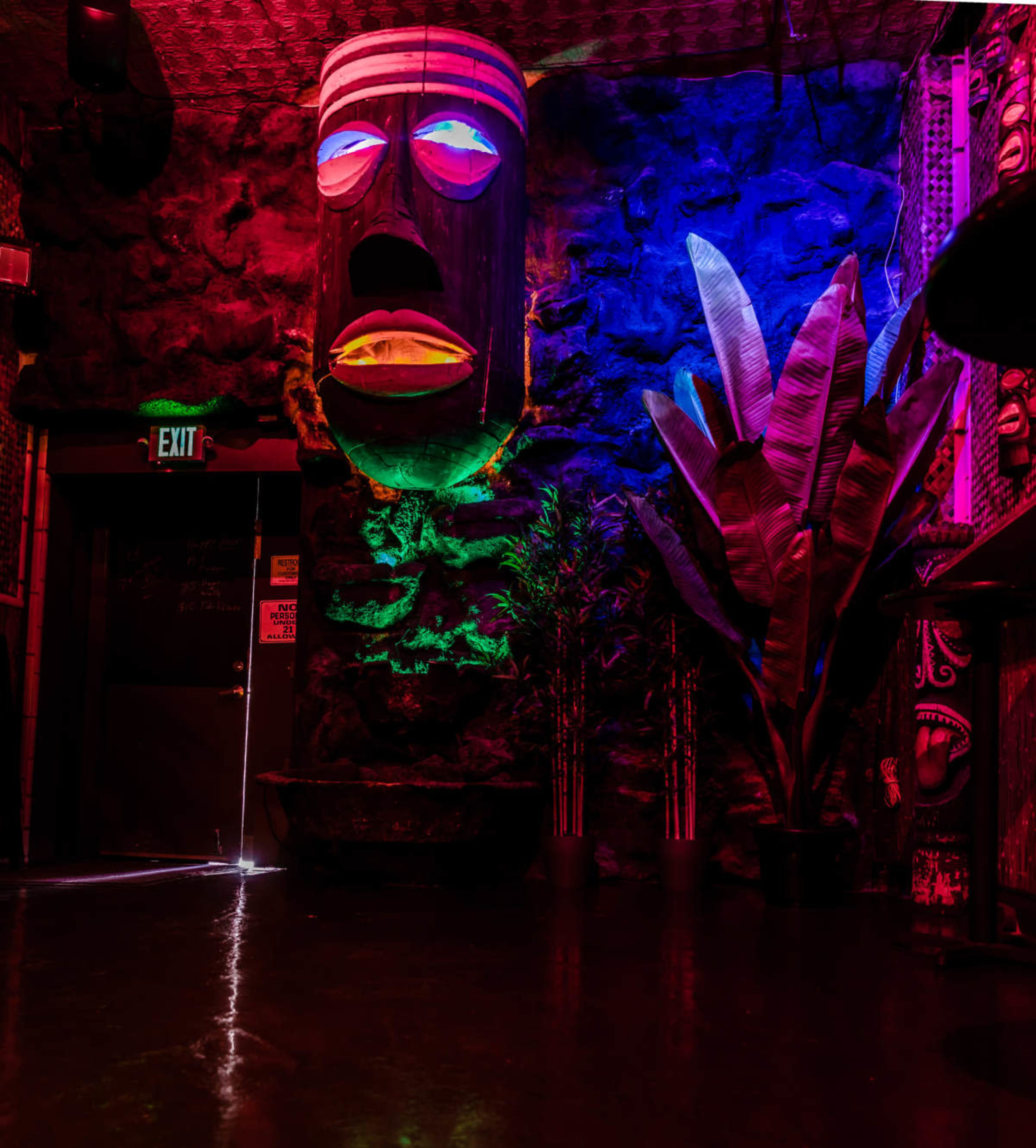 The image features a dimly lit room with a large, colorful tiki mask on a stone wall, accompanied by tropical plants and an exit door.