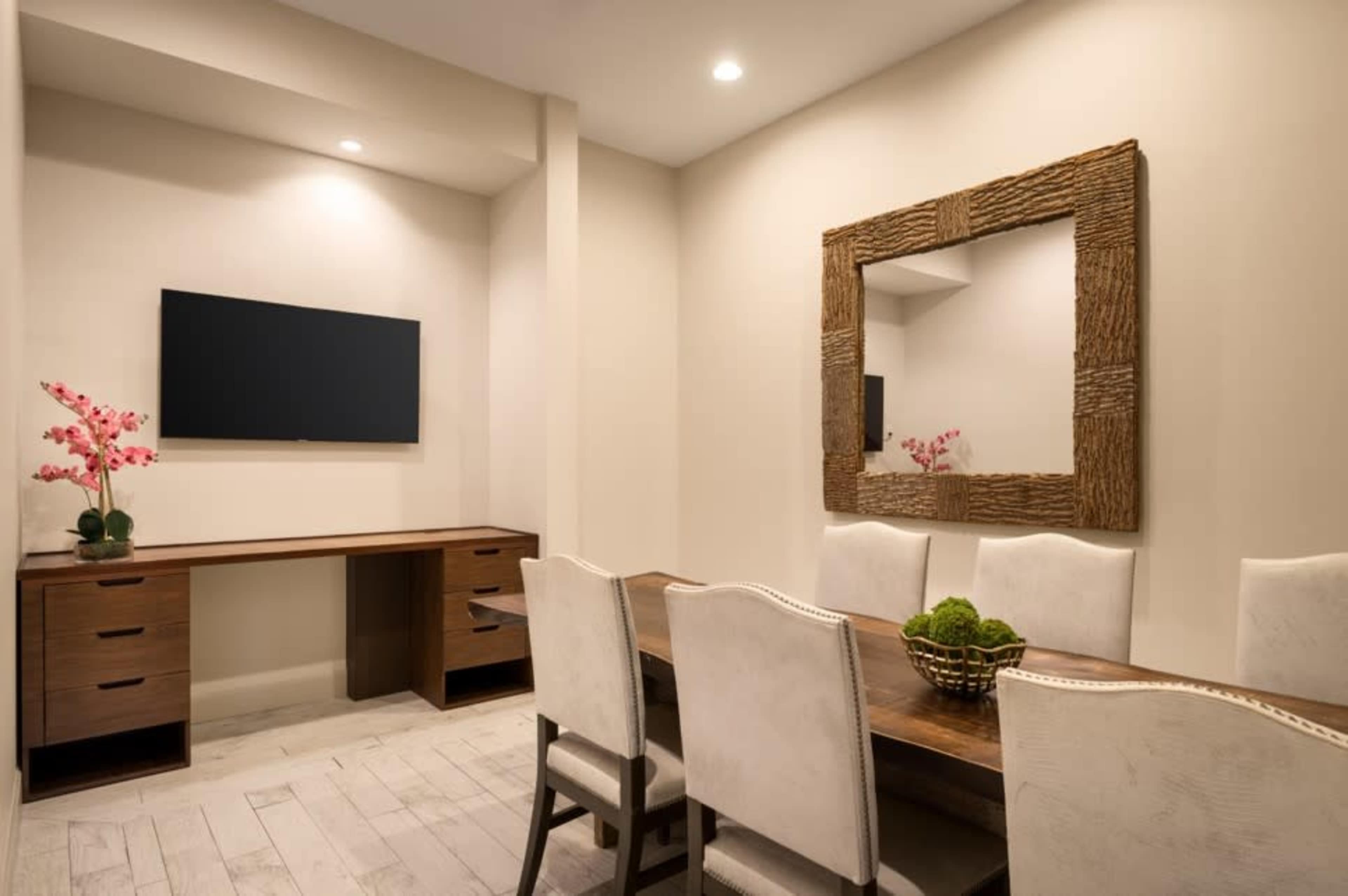 A modern dining room features a long wooden table, upholstered chairs, a wall-mounted television, and a decorative mirror, with floral arrangements and greenery accentuating the space.