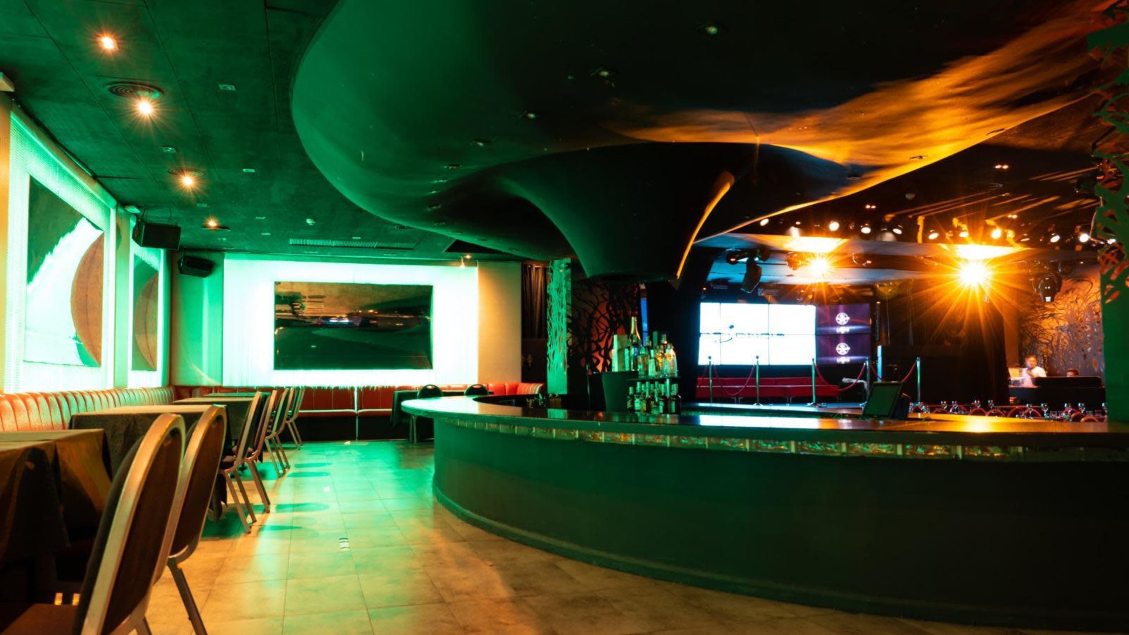 The image shows a modern nightclub interior with a curved bar, stage lighting, and booths arranged around the dance floor.