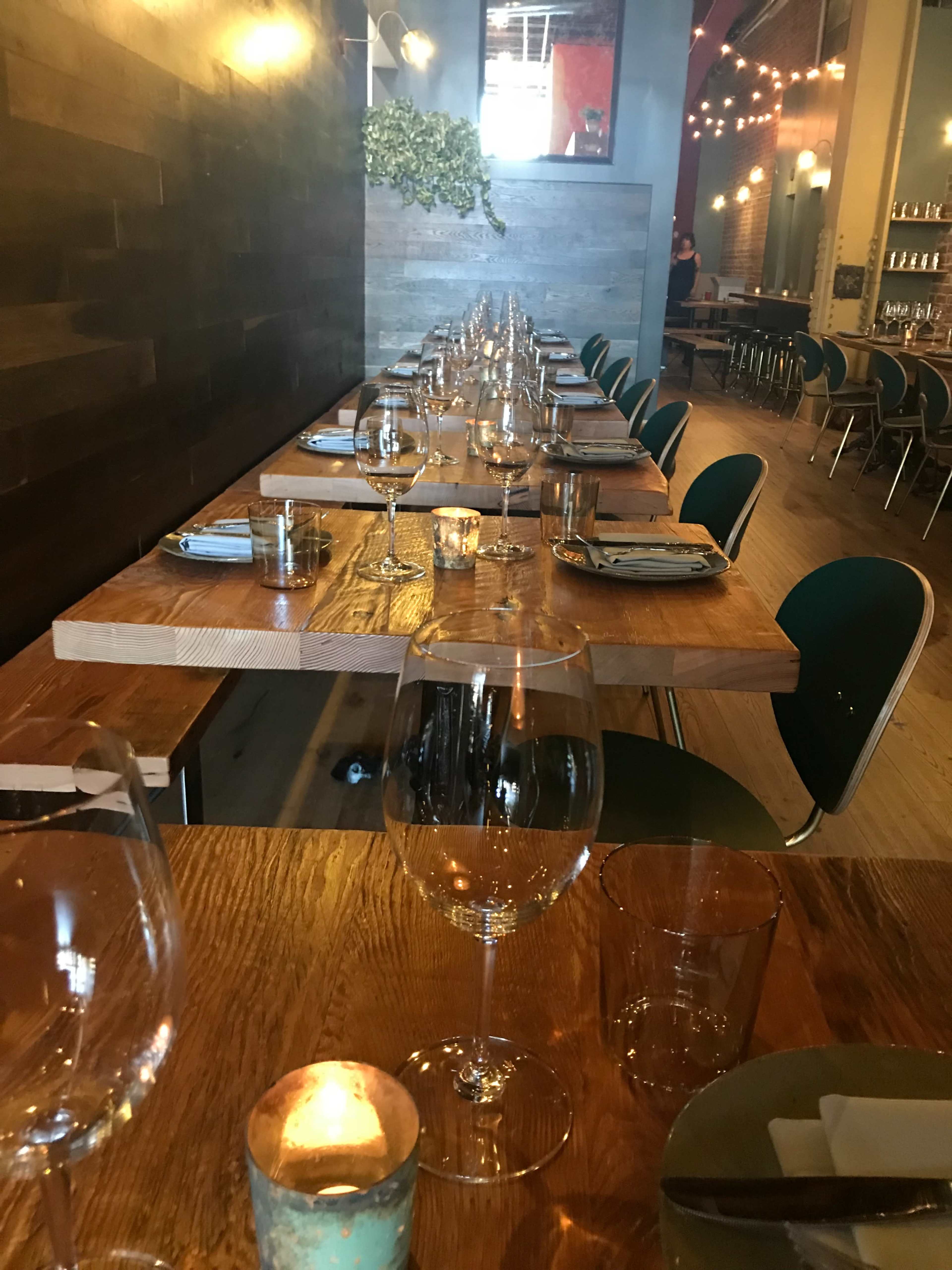 A long wooden dining table is set with wine glasses and candles in a restaurant interior.