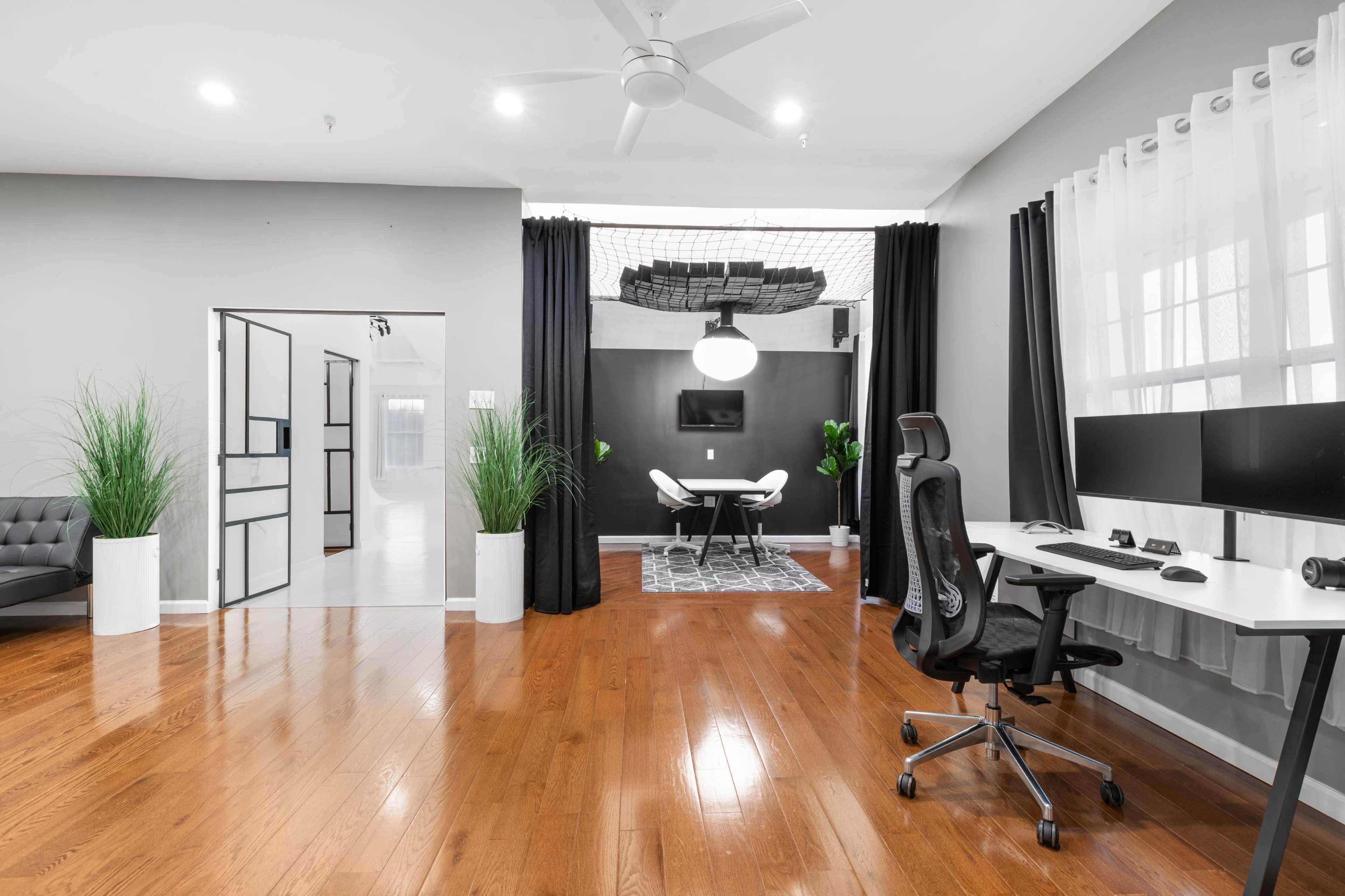 A modern home office with a sleek desk, two monitors, and a seating area, surrounded by plants and a contrasting dark backdrop.
