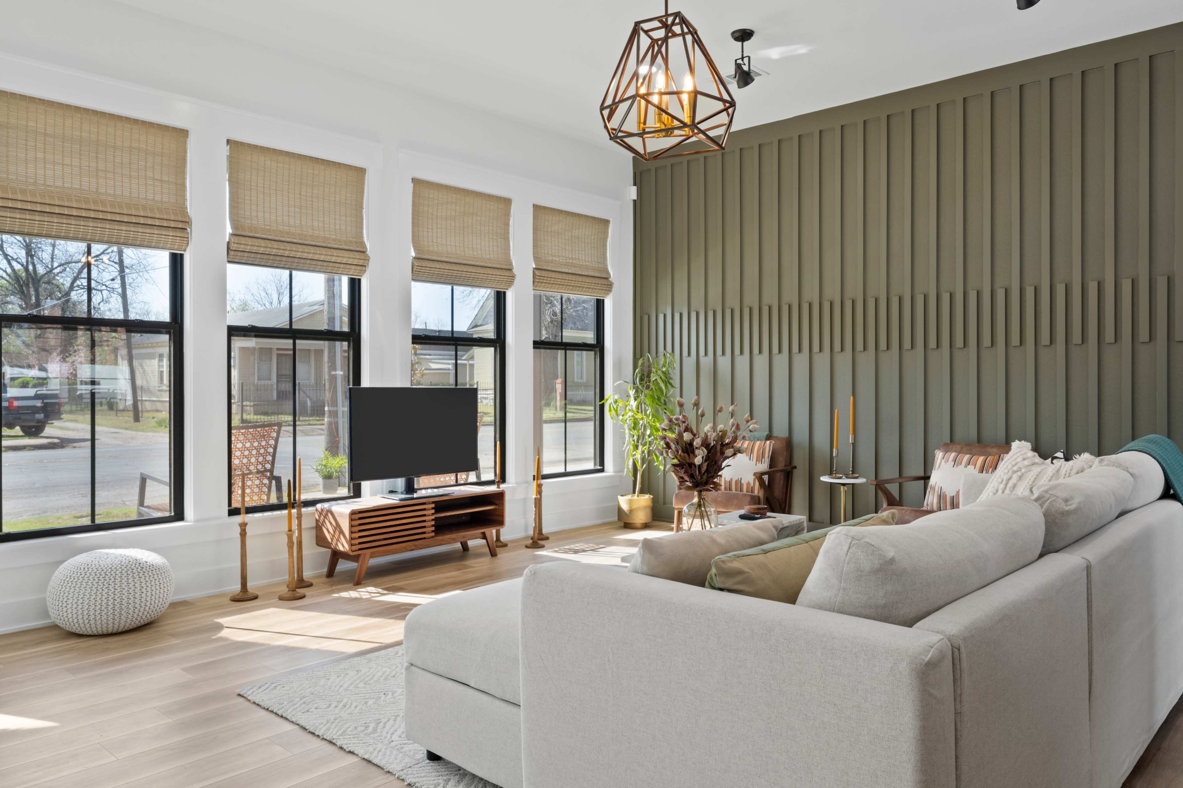 A modern living room features a sectional sofa, a wooden TV stand with a television, and large windows adorned with woven shades, all set against a green accent wall.