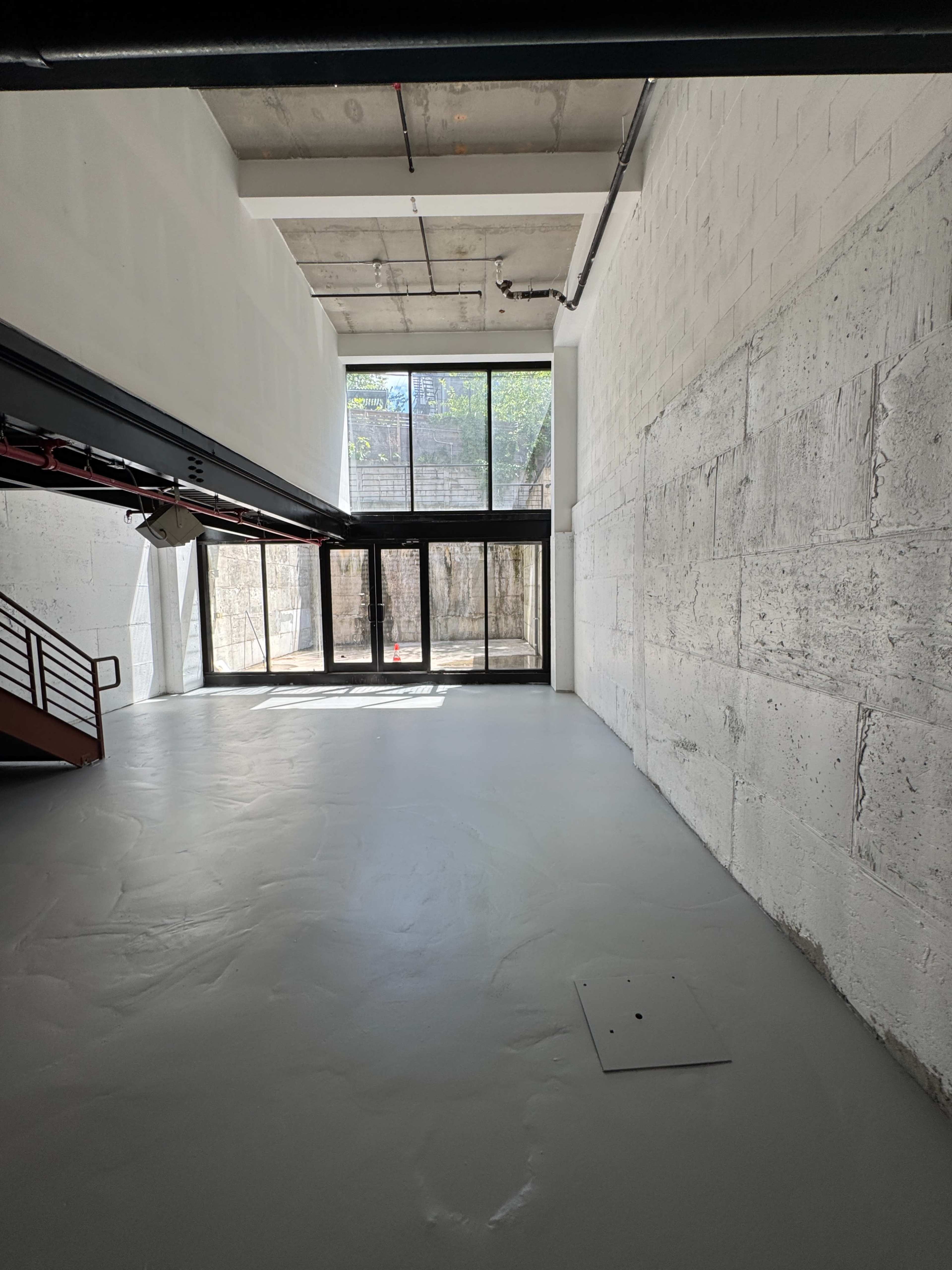 The image shows a spacious, empty interior with high ceilings, large windows, and a concrete wall.