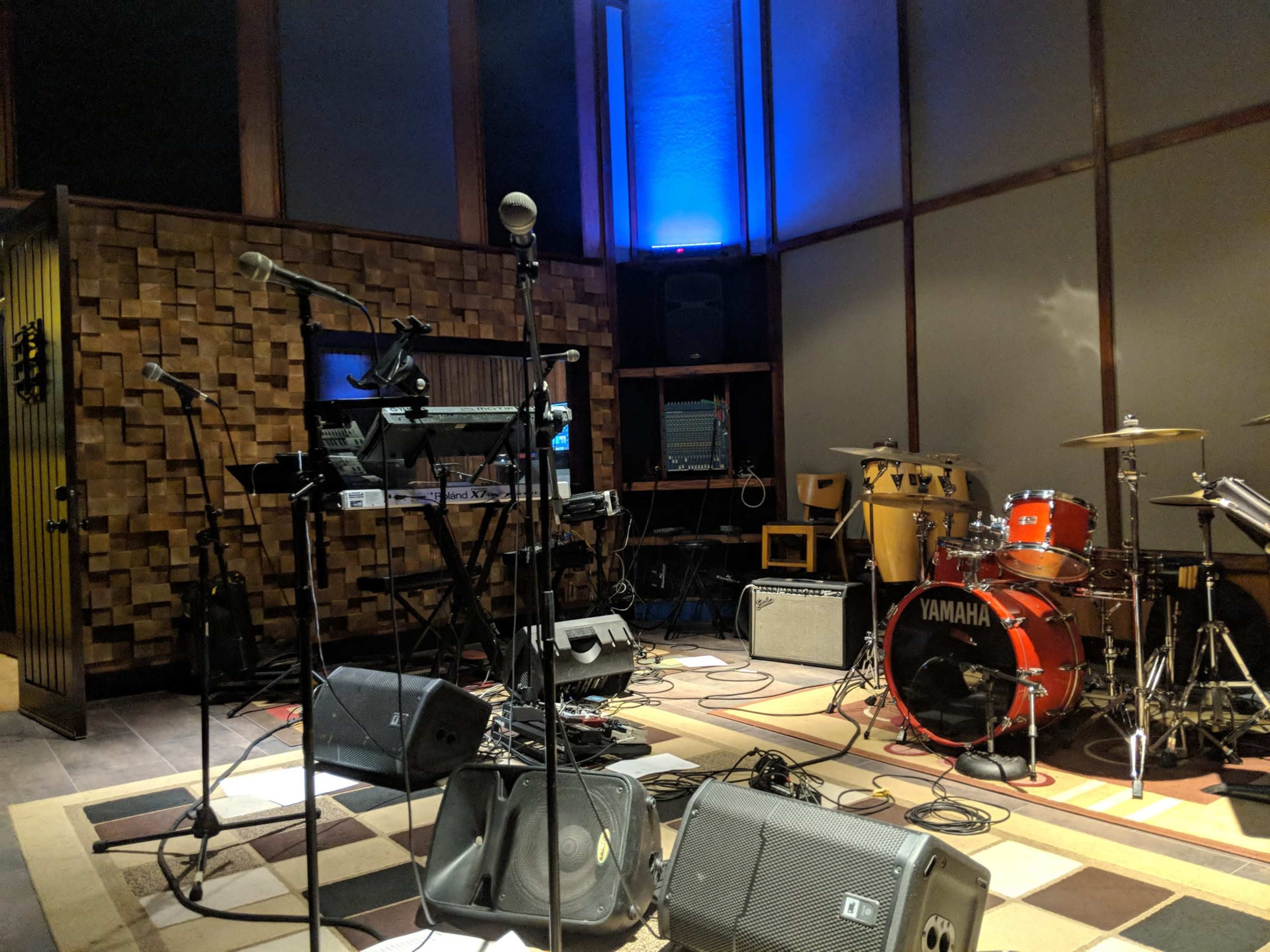 A music rehearsal room equipped with various instruments, including microphones, drums, keyboards, and amplifiers, arranged on a carpeted floor.