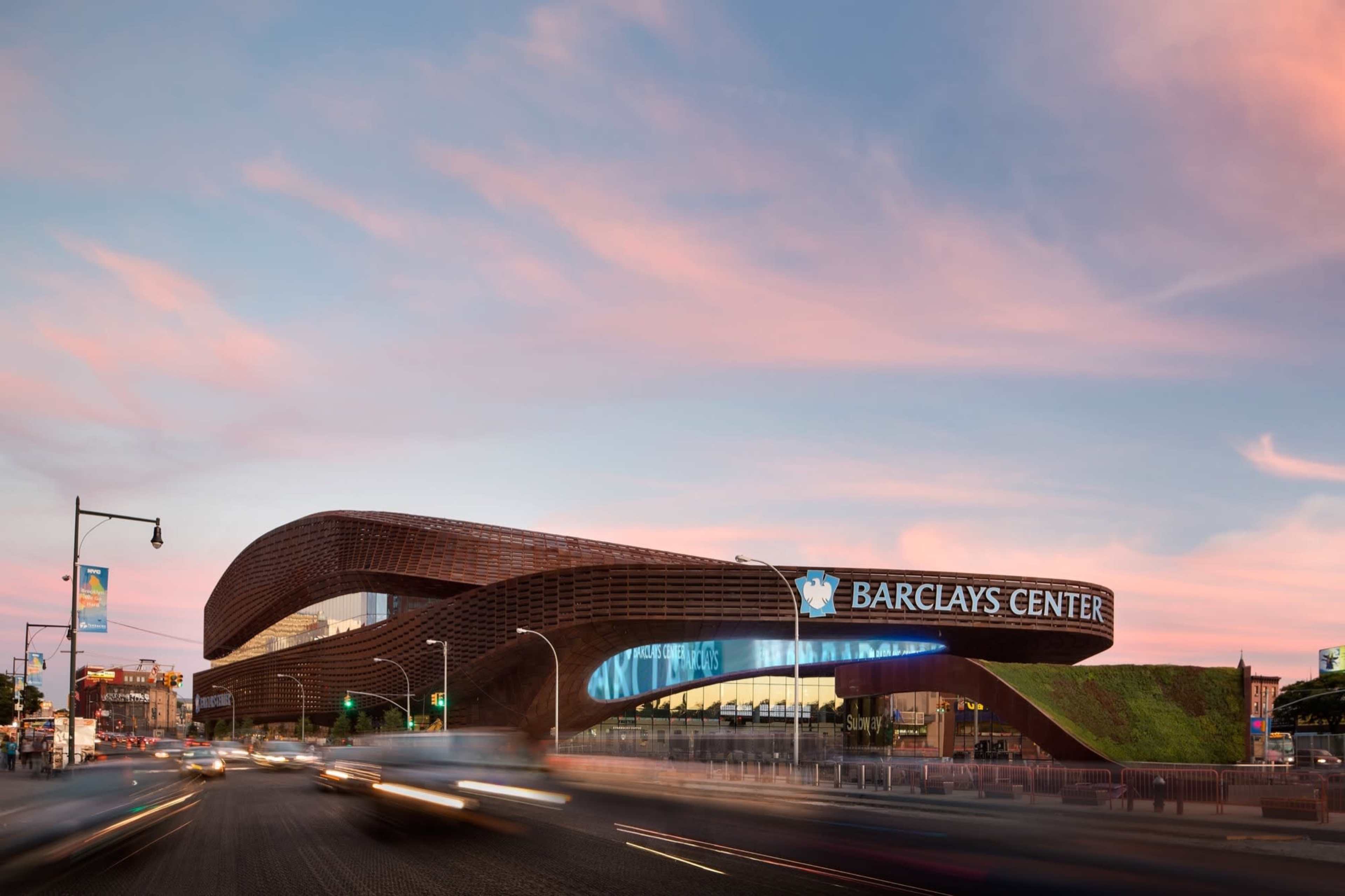 The image shows the Barclays Center, a sports and entertainment arena in Brooklyn, New York, featuring a distinctive curved exterior design and illuminated signage.