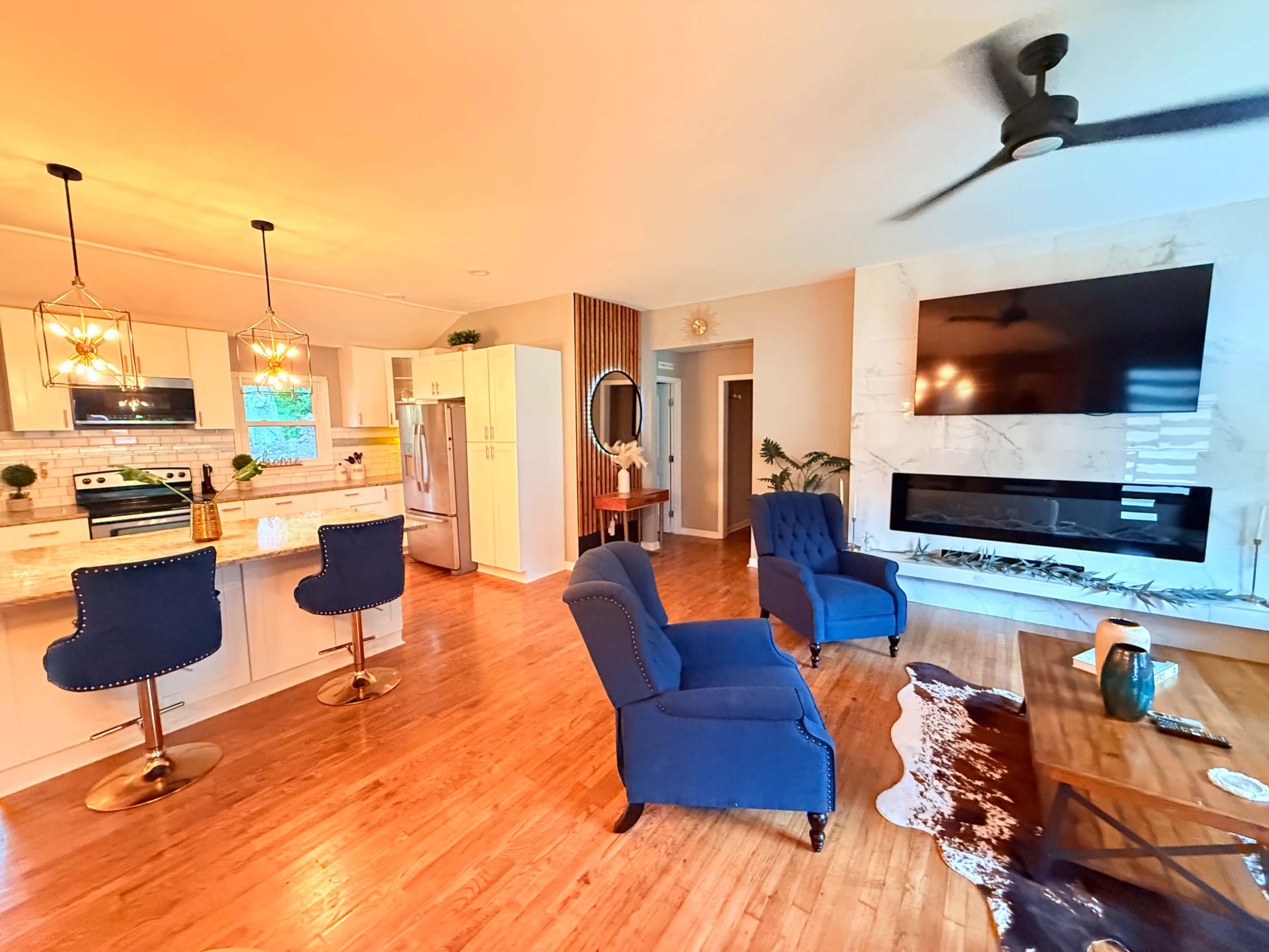 The image shows a modern kitchen and living area featuring blue upholstered chairs, a wooden floor, a large wall-mounted TV, and a sleek kitchen with white cabinetry and stainless steel appliances.