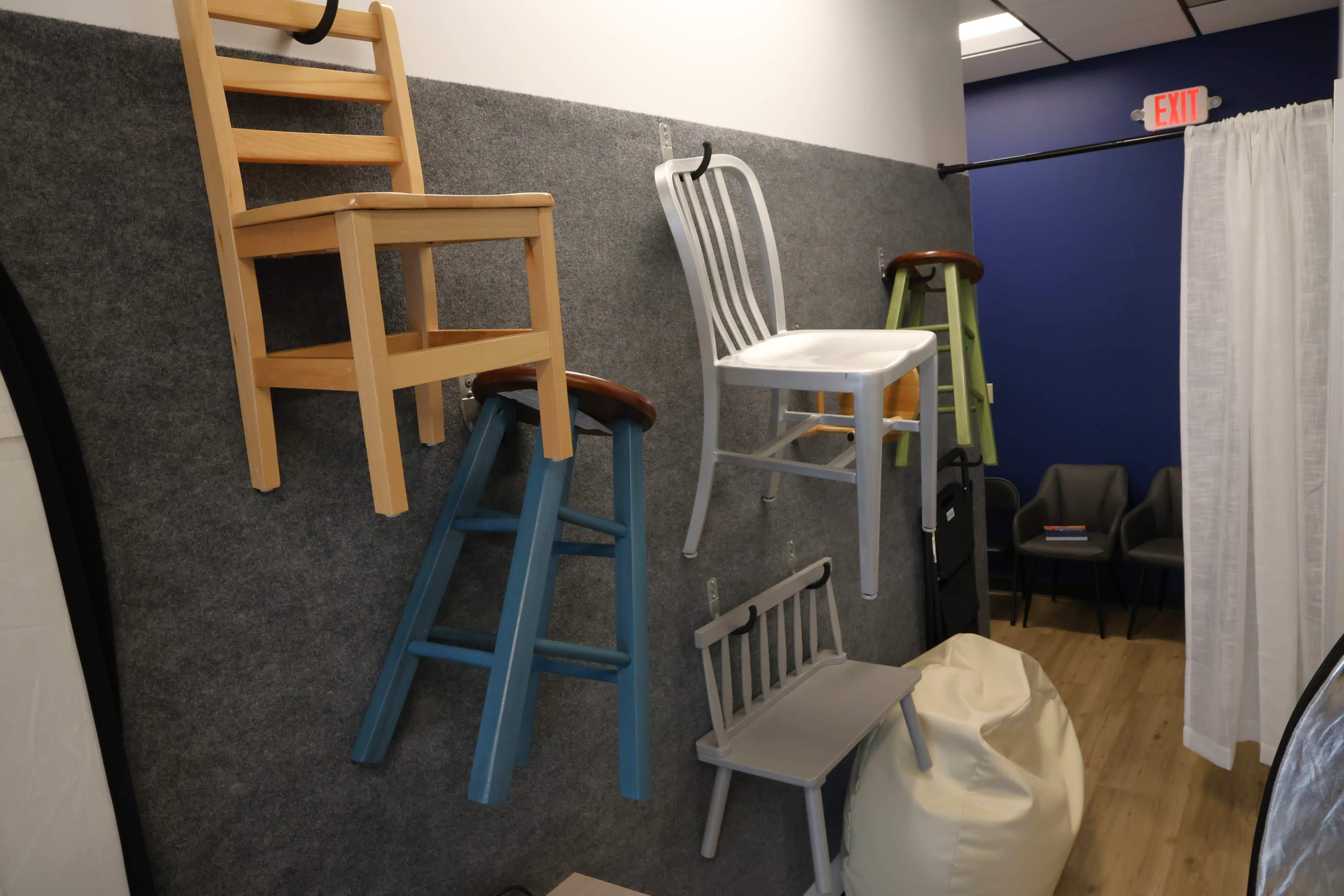 Several colorful chairs are mounted on a gray felt wall in an indoor space.