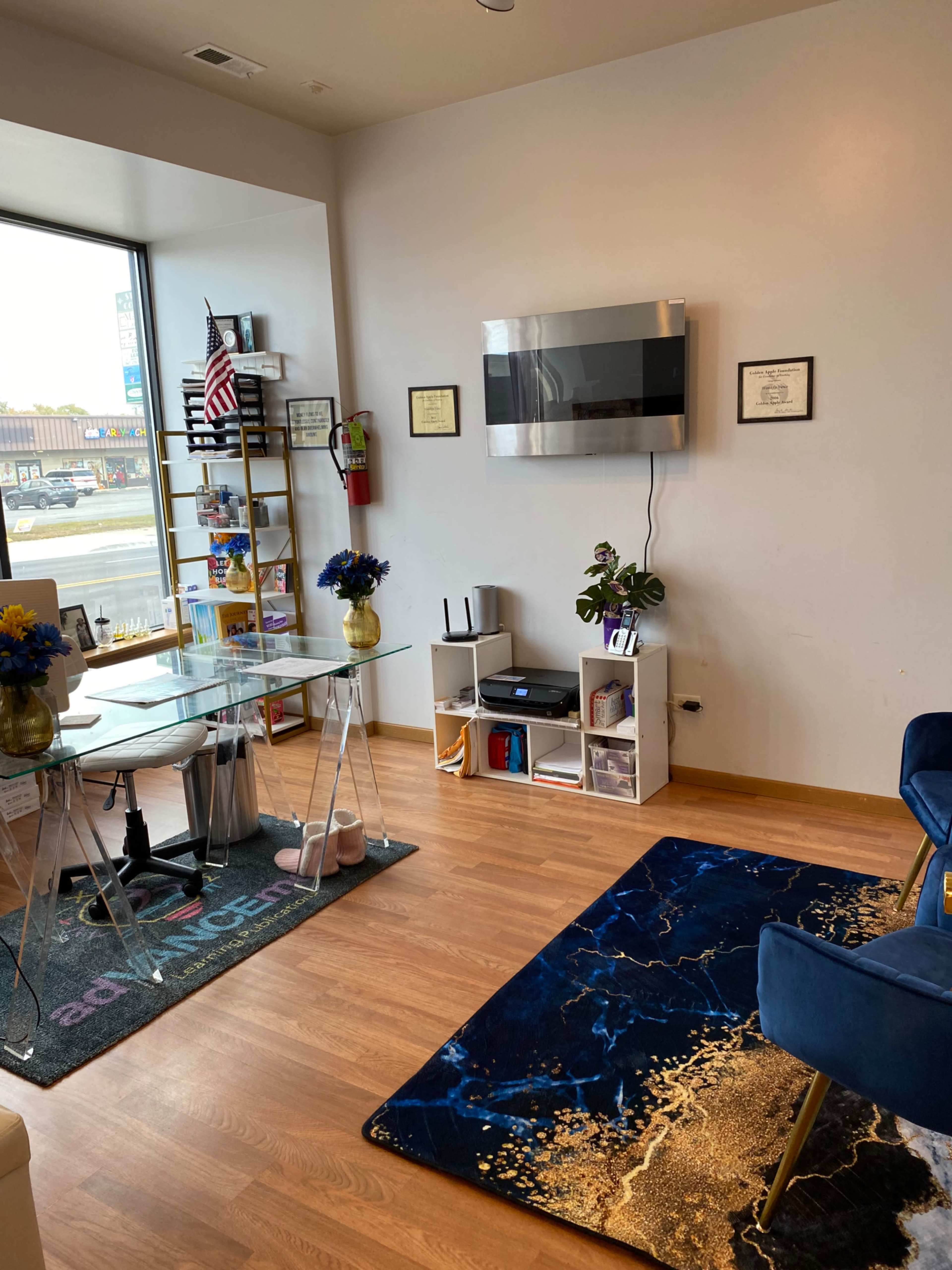 An office space featuring a glass dining table with chairs, a wall-mounted television, certificates framed on the wall, and a blue rug on the wooden floor.