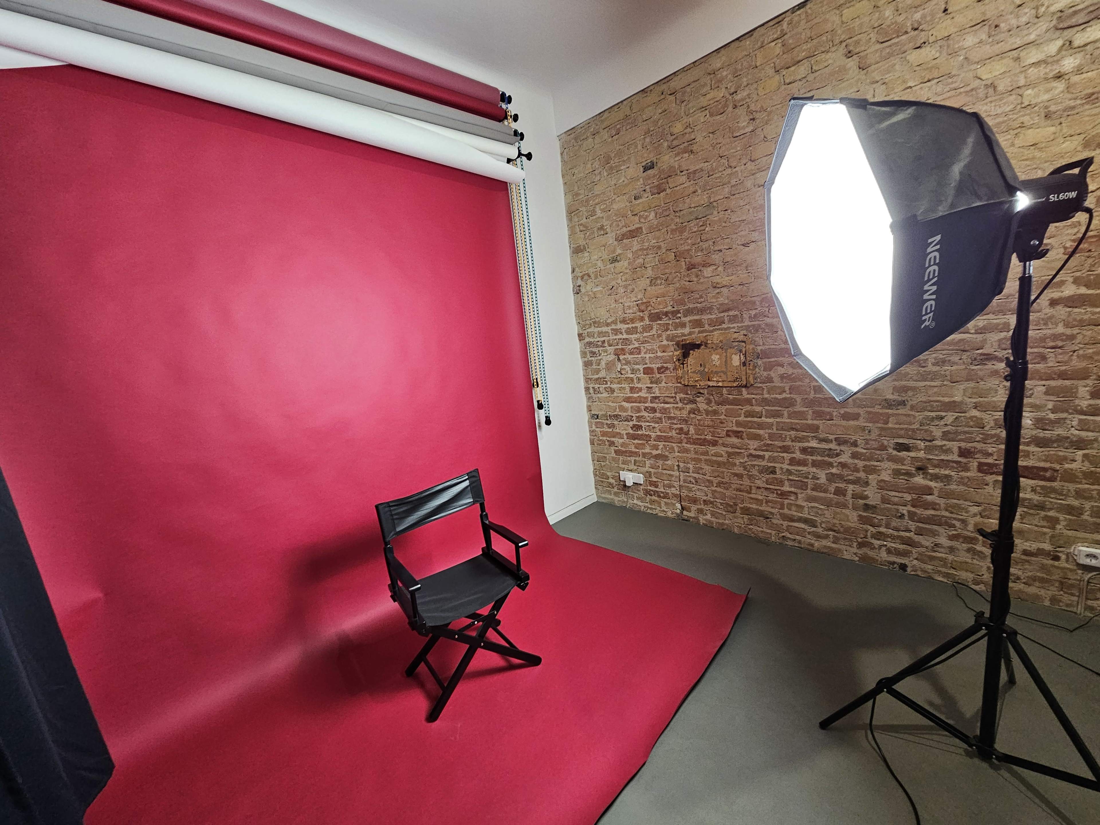 The image shows a photography studio with a bright red backdrop, a black director's chair, and a large softbox light.