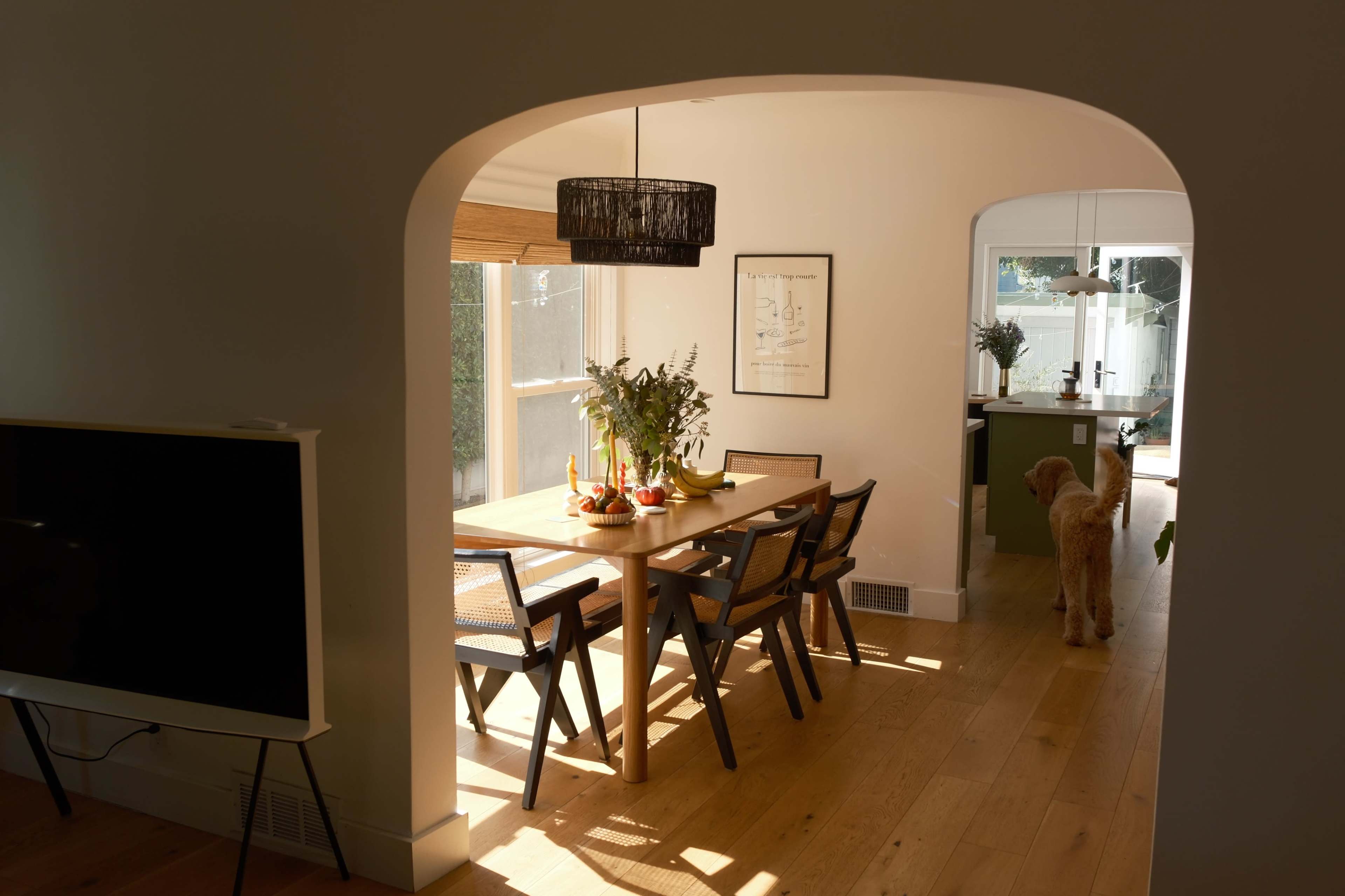 A dining area features a wooden table surrounded by chairs, with a large window allowing natural light to enter, and a dog standing nearby.