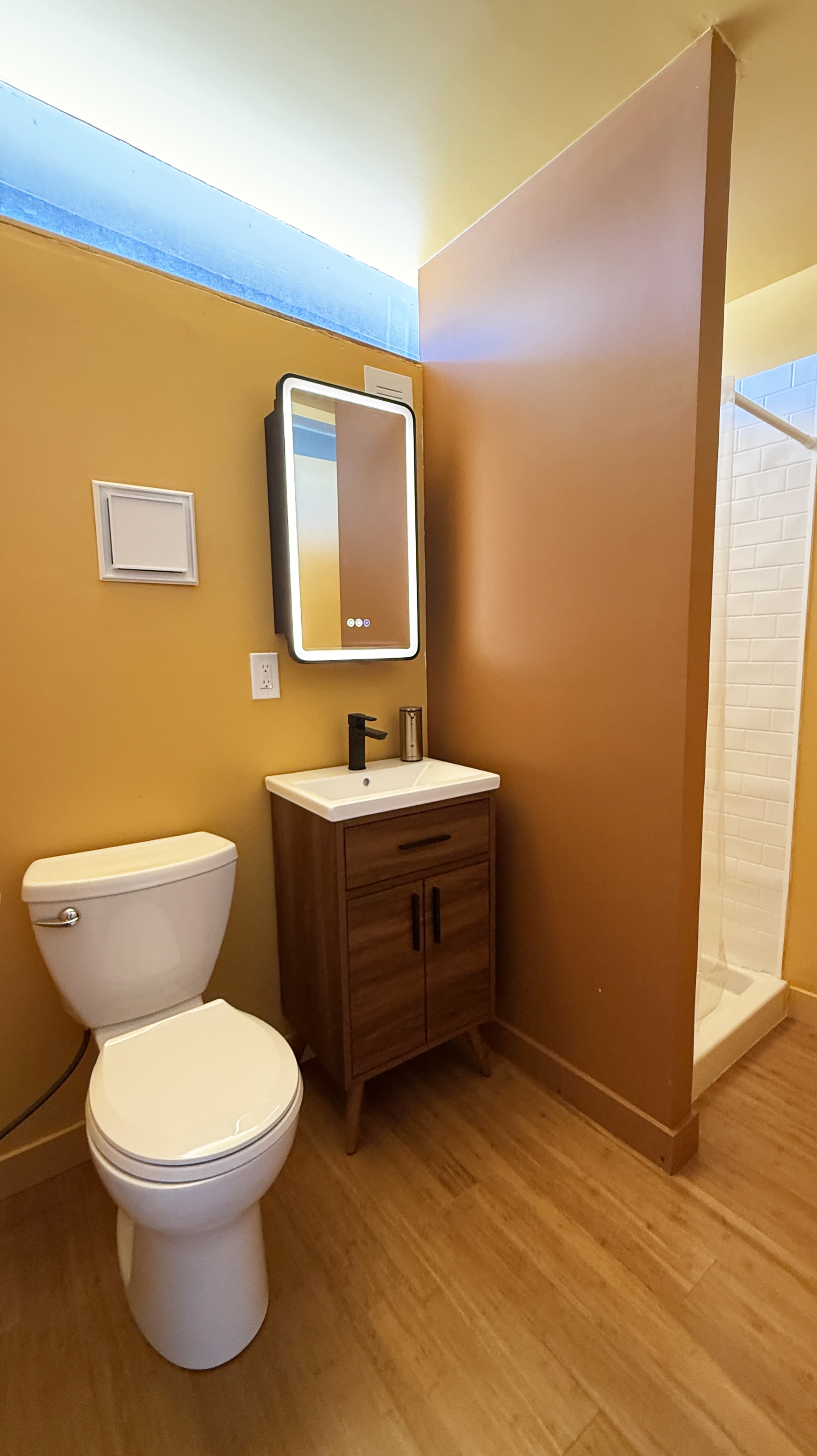 The image shows a small bathroom featuring a toilet, a wooden vanity with a sink, a mirror, and a shower stall, all set against yellow walls.