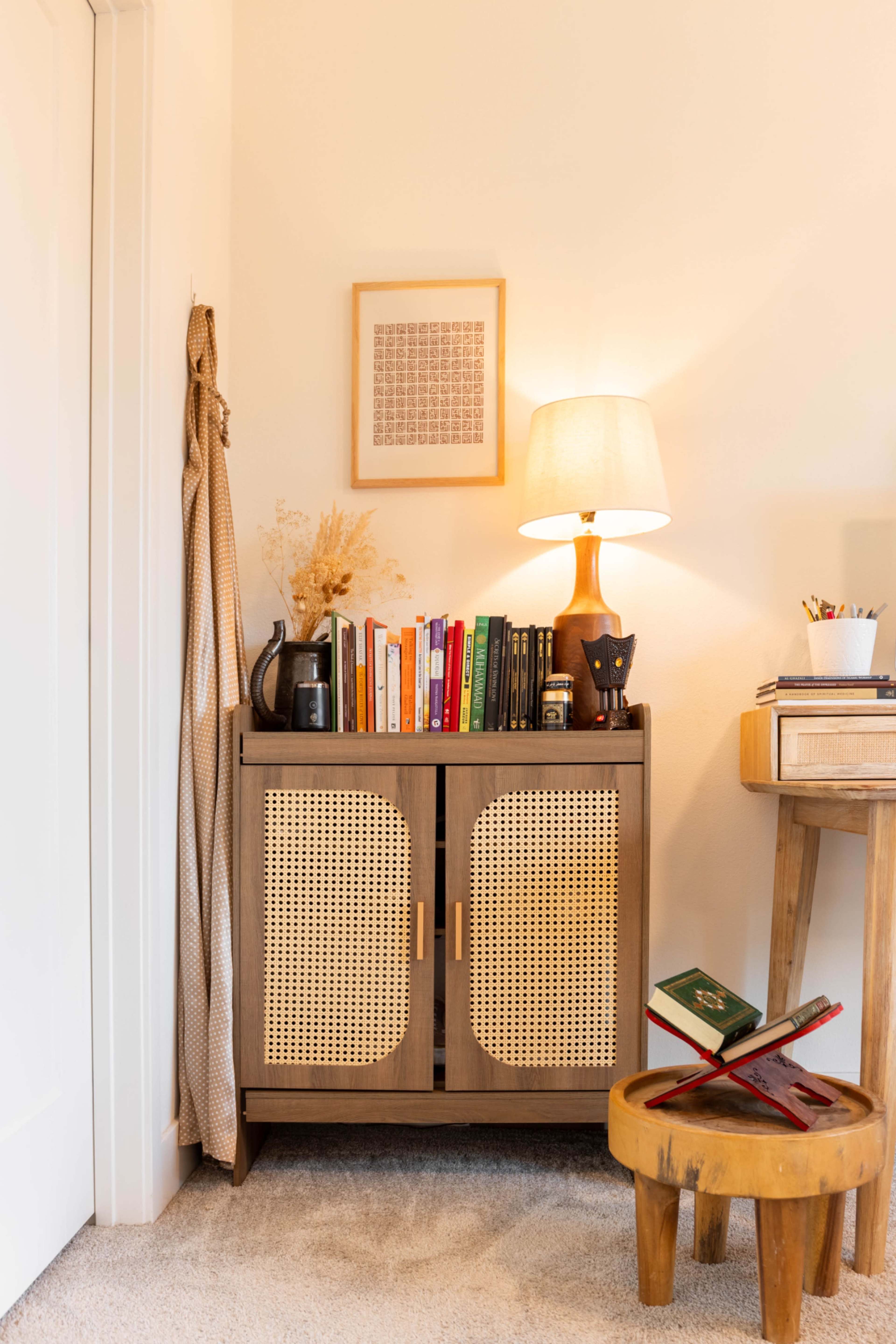 The image shows a corner of a room featuring a bookshelf with closed doors, a table lamp on top, a small round stool, and a framed piece of art on the wall.
