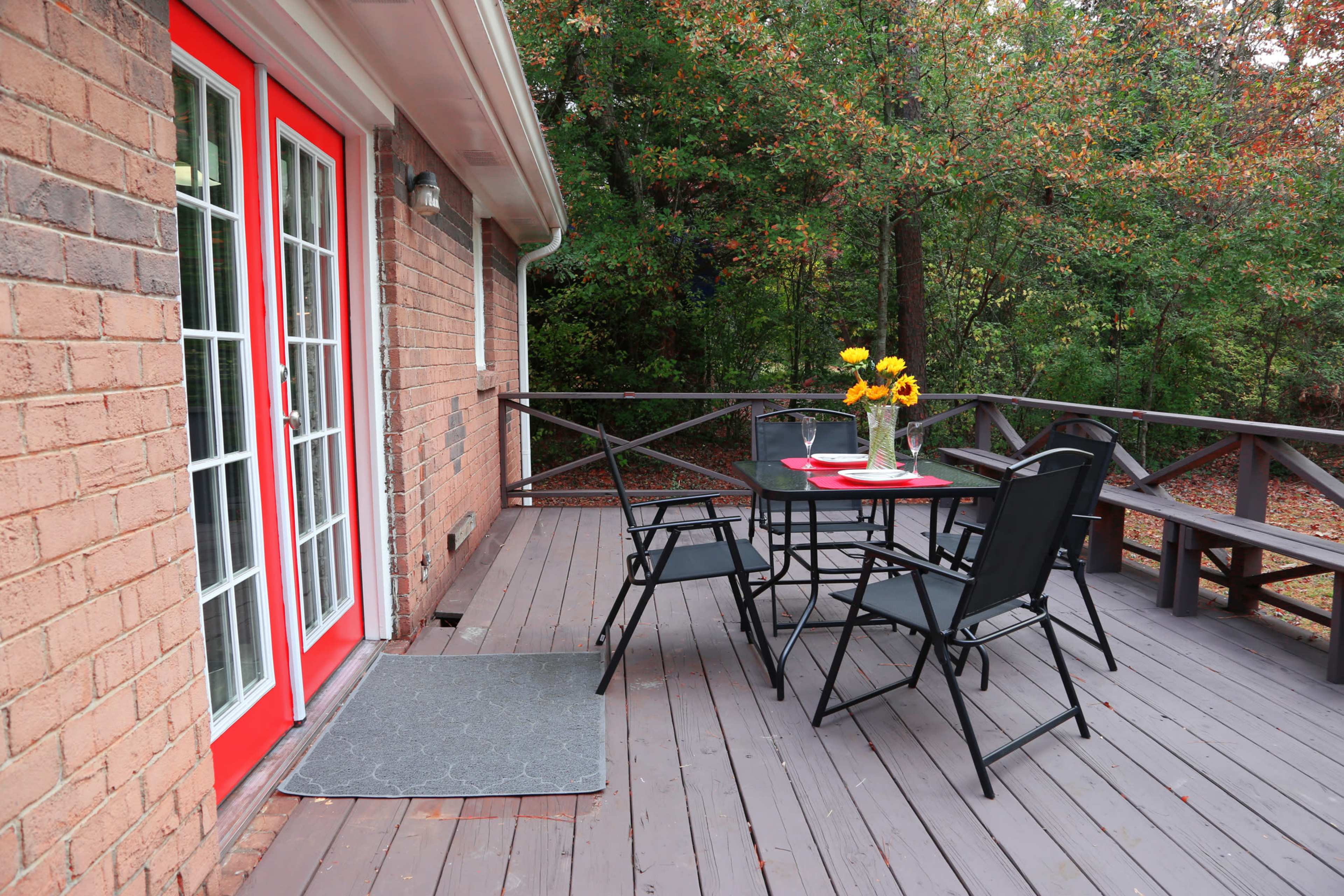 A wooden deck with a table and four chairs, featuring a vase of sunflowers, alongside a brick house with red French doors.