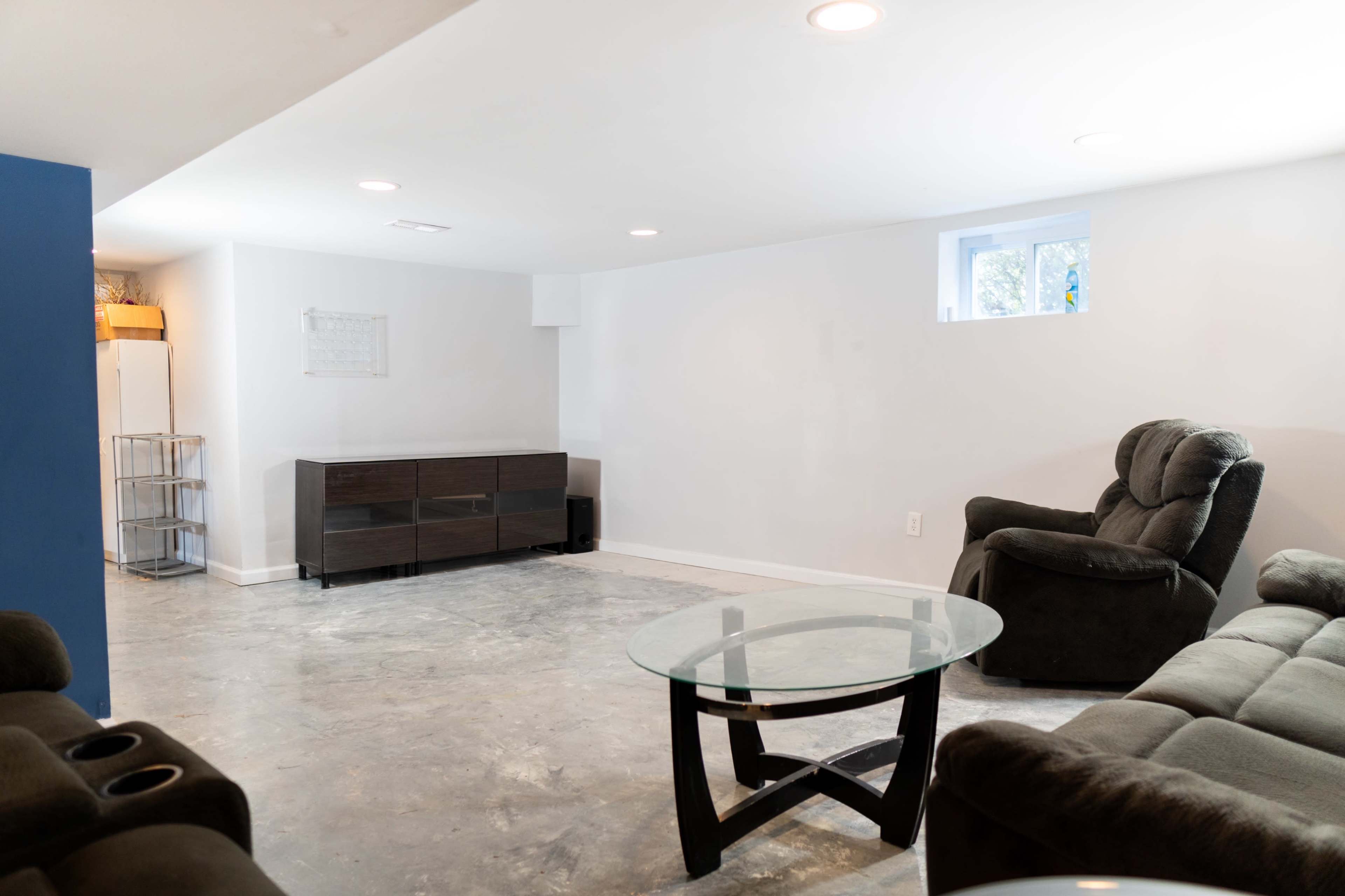 The image shows a sparsely furnished basement with a glass coffee table, two couches, and a small cabinet against a plain wall.