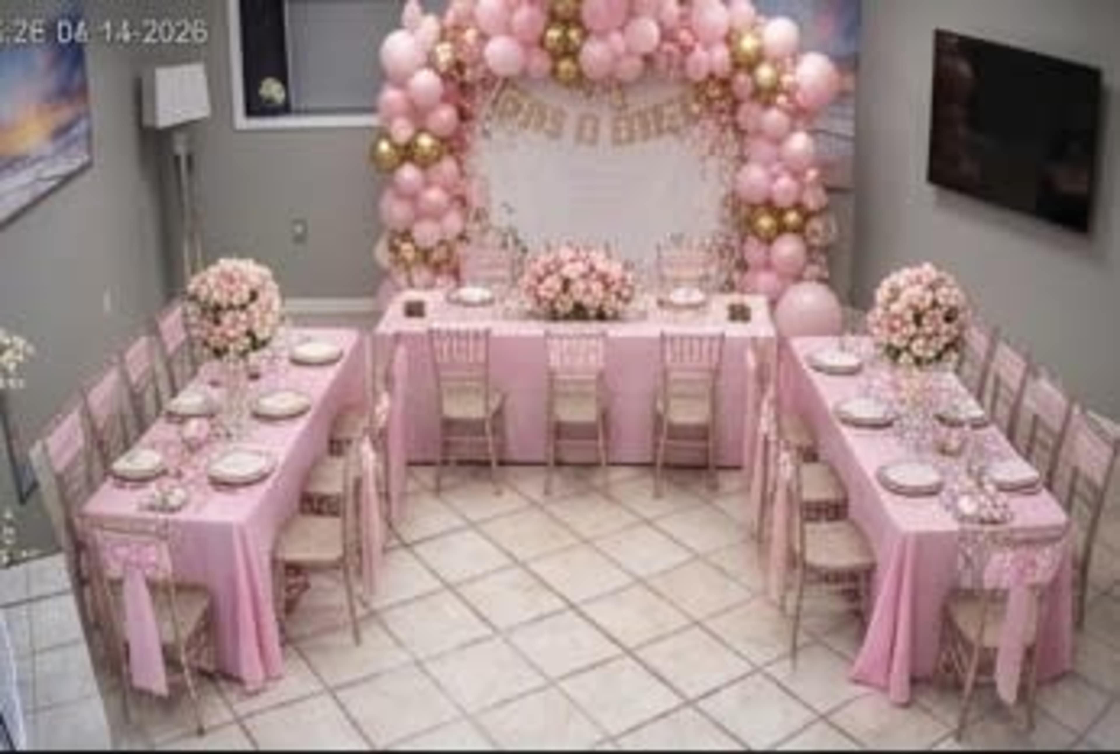 The image shows a decorated event space featuring two long tables covered in pink tablecloths, surrounded by chairs, with balloon arches and floral arrangements in a light pastel color scheme.