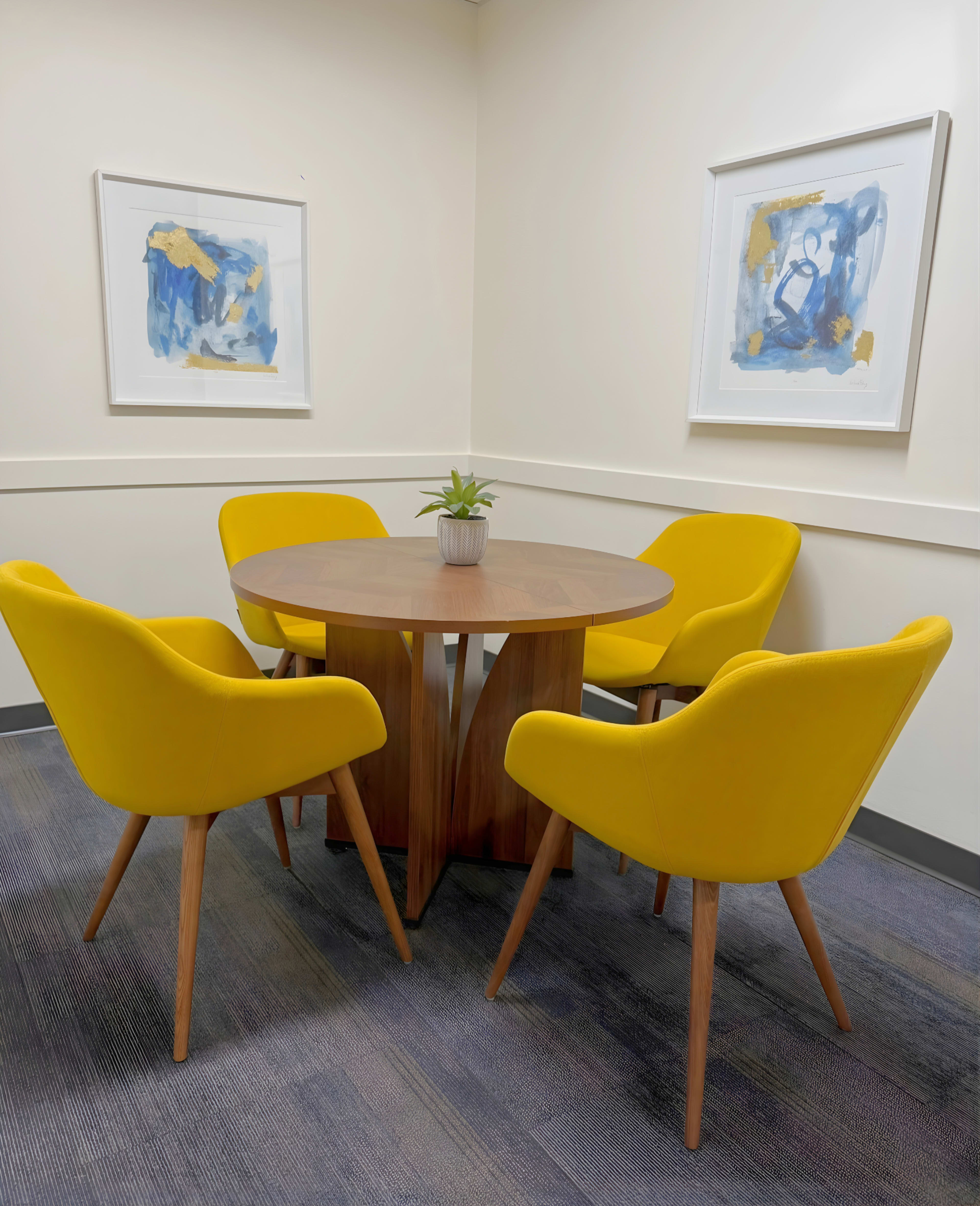 A round wooden table surrounded by four yellow chairs sits in a corner of a room, with two framed abstract paintings on the walls.