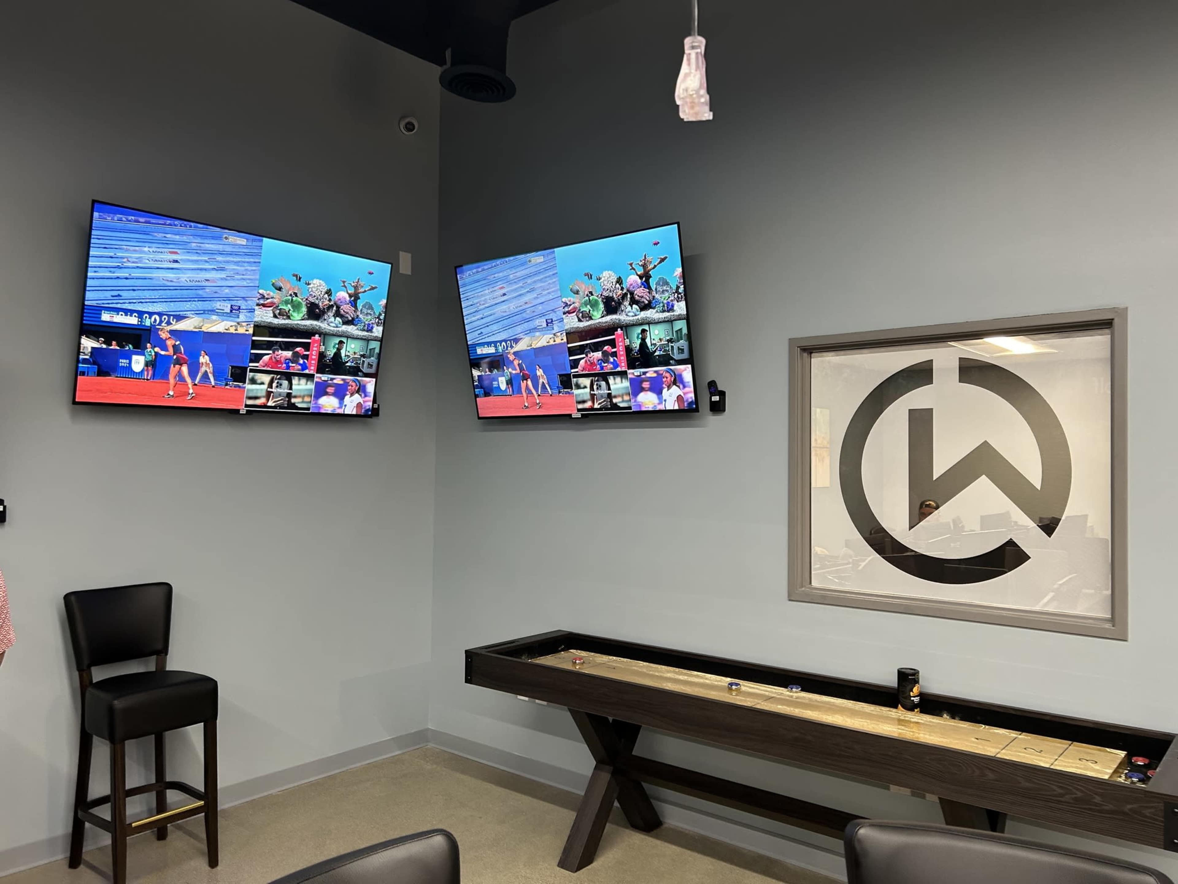 A modern room with two mounted televisions displaying a sports event and a shuffleboard table on one side, along with a logo on the wall.