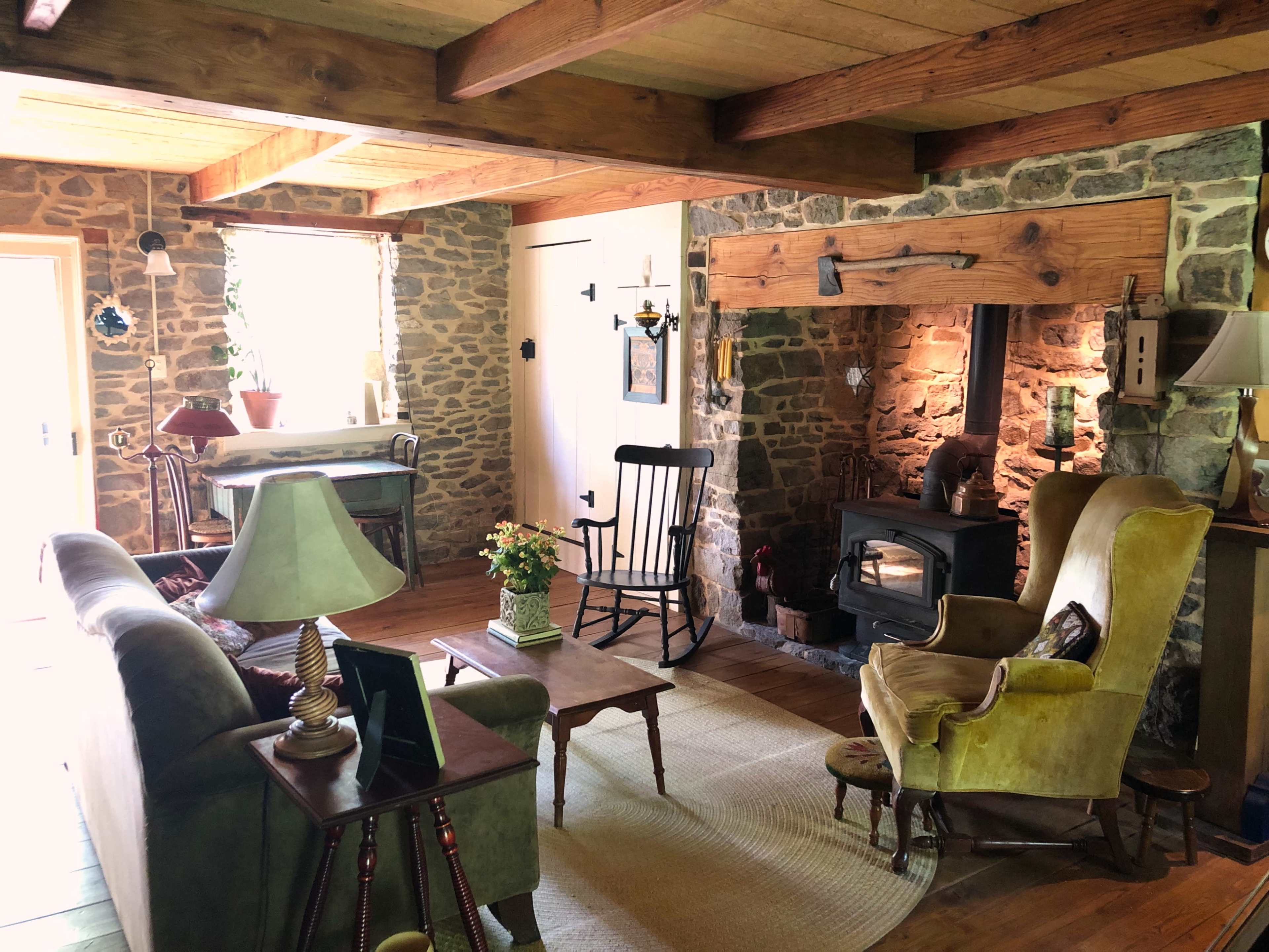 The image shows a cozy living room featuring stone walls, wooden beams, a fireplace, and a mix of vintage furniture including a rocking chair and upholstered armchairs.