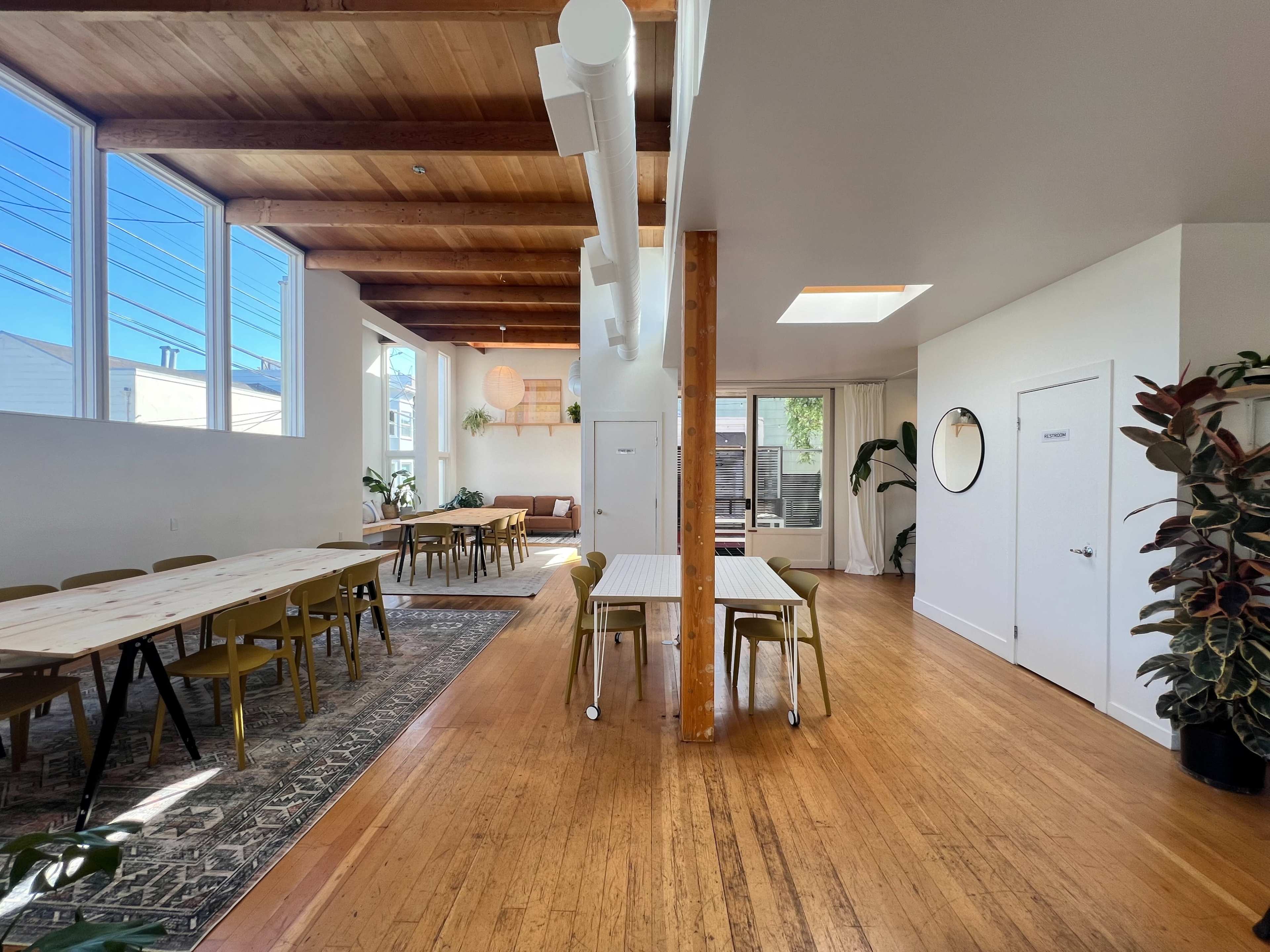 The image shows a spacious, brightly lit interior with wooden beams, tables arranged for gatherings, and plants along the walls.