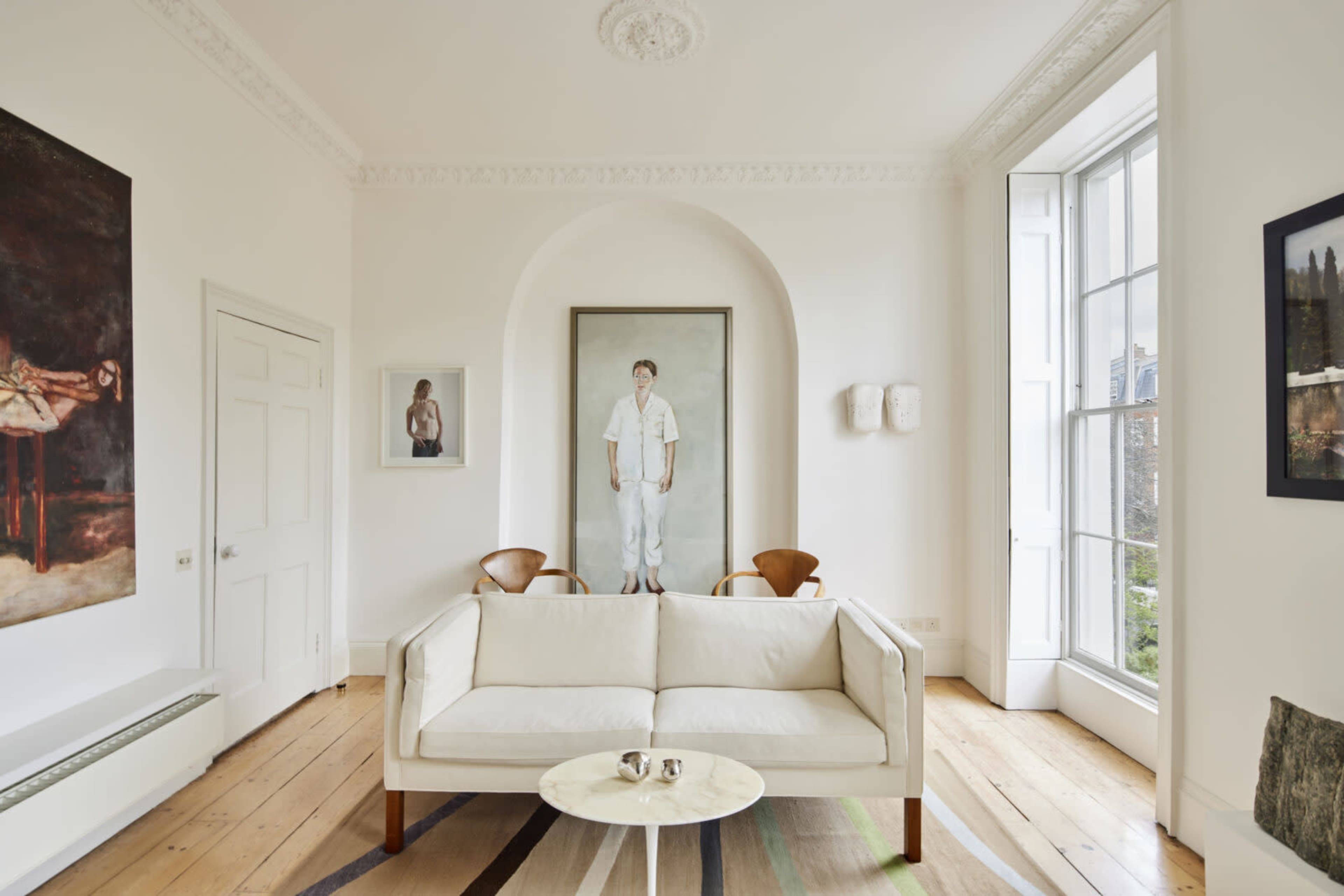 A bright living room featuring a white sofa, two wooden chairs, and framed artworks on the walls.