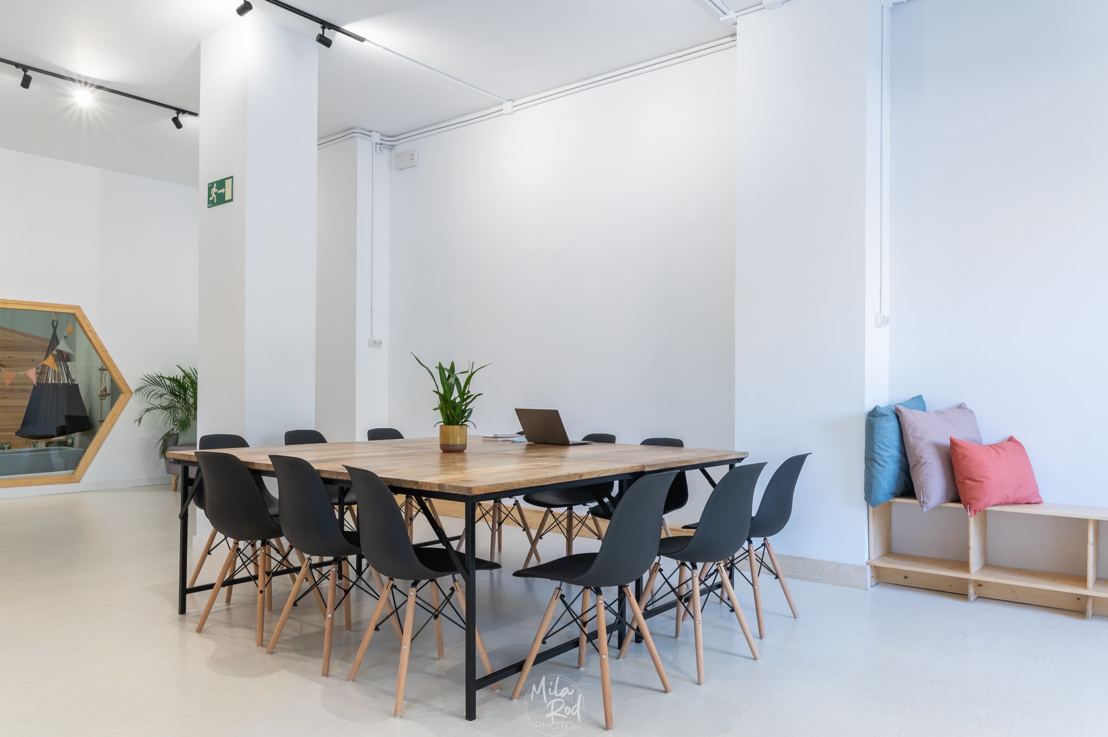 A large wooden table surrounded by black chairs is set up in a bright room with white walls and a few decorative elements.