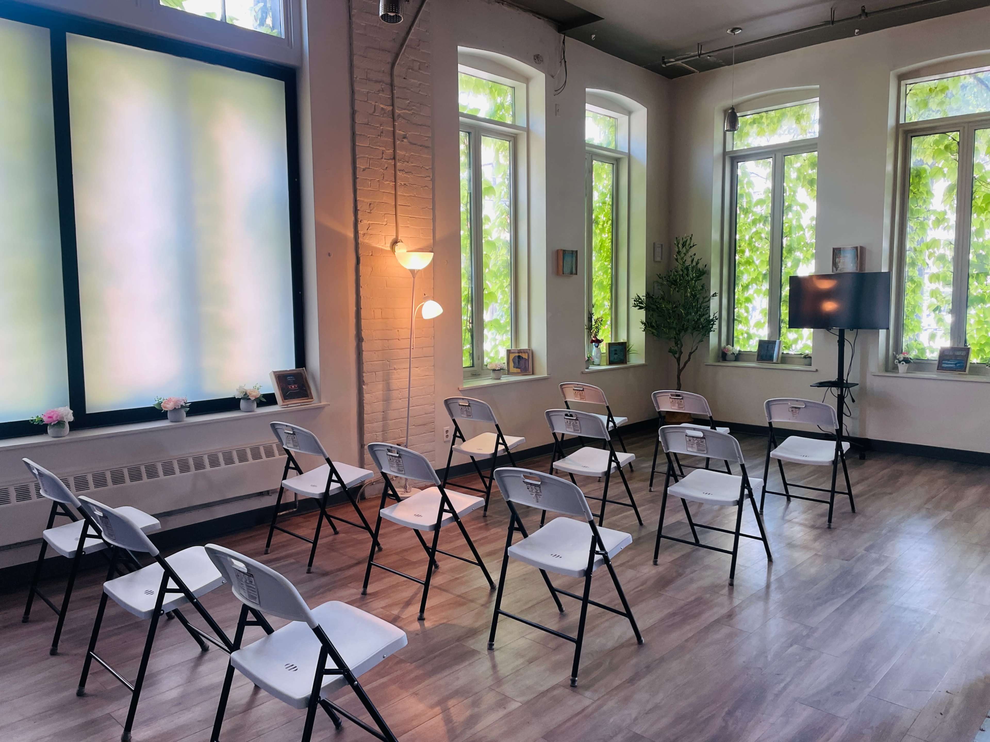 A small, bright room arranged with white folding chairs facing a TV, surrounded by large windows with greenery outside.