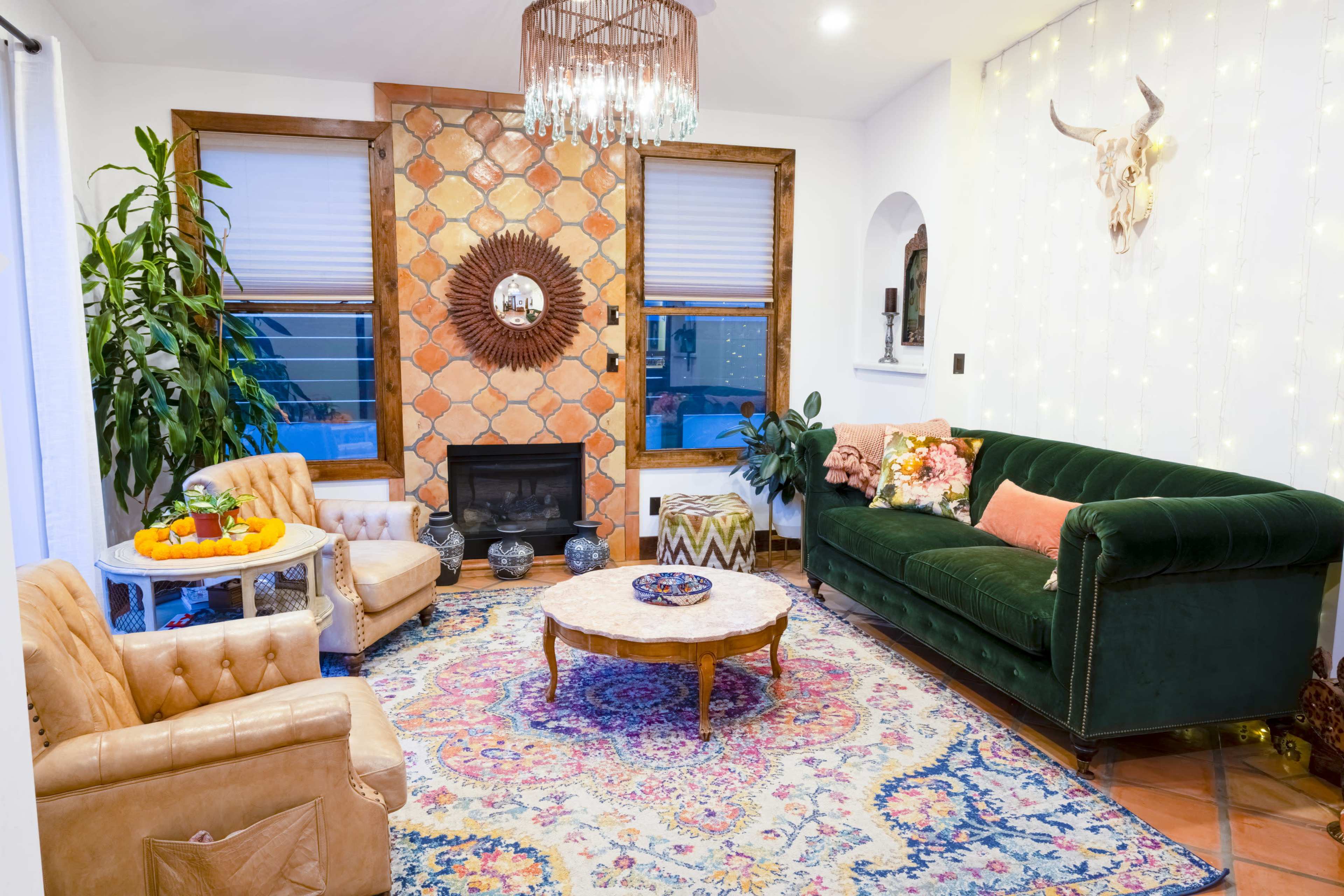 The living room features a green velvet sofa, a round wooden coffee table, two armchairs, a patterned area rug, and a decorative wall with a mounted animal head and a sunburst mirror.