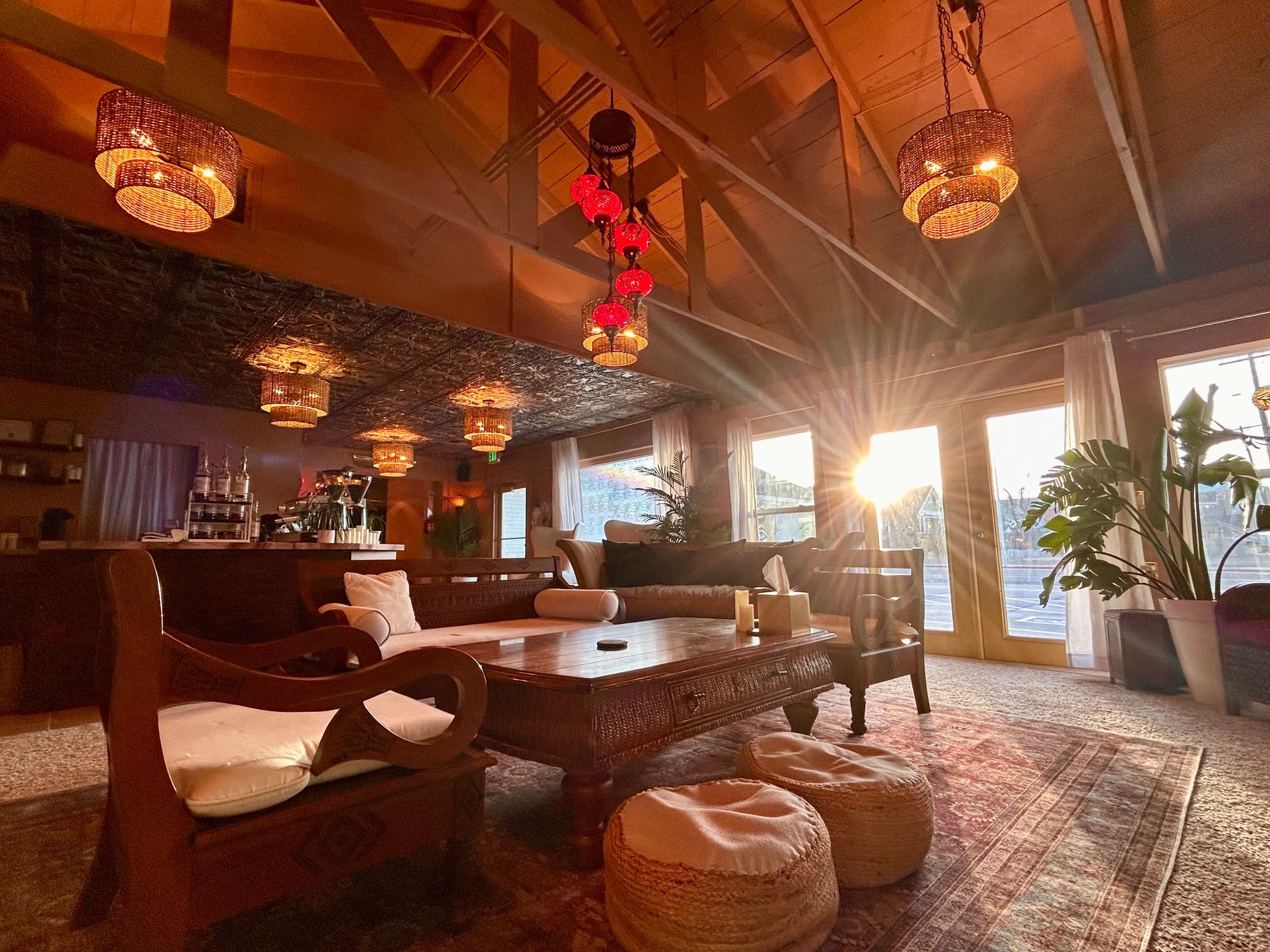 A warm, inviting living room features wooden beams, wicker pendant lights, a large wooden coffee table, and a view of the sunset through glass doors.