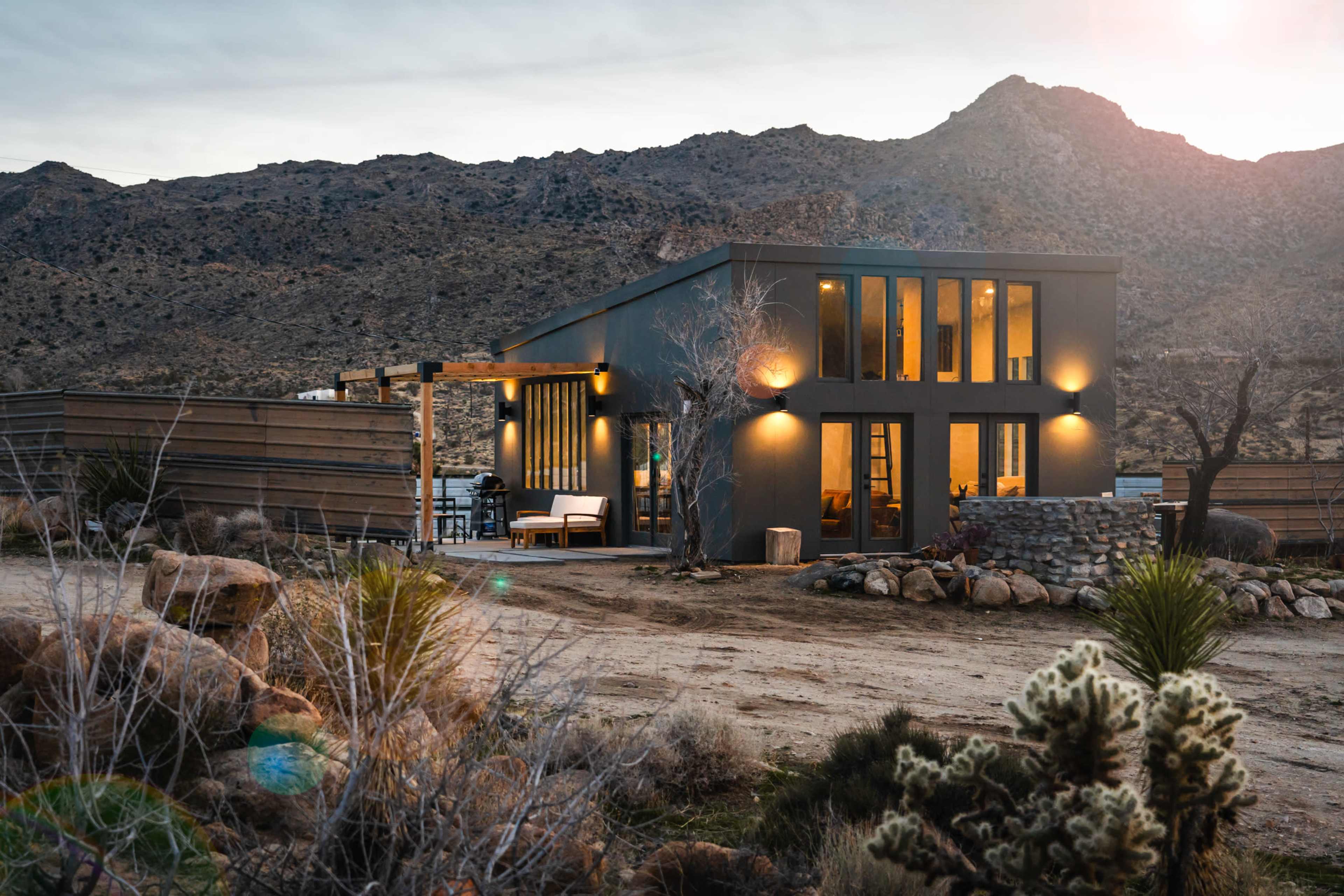 A modern house with large windows sits in a desert landscape, surrounded by rocky terrain and sparse vegetation.