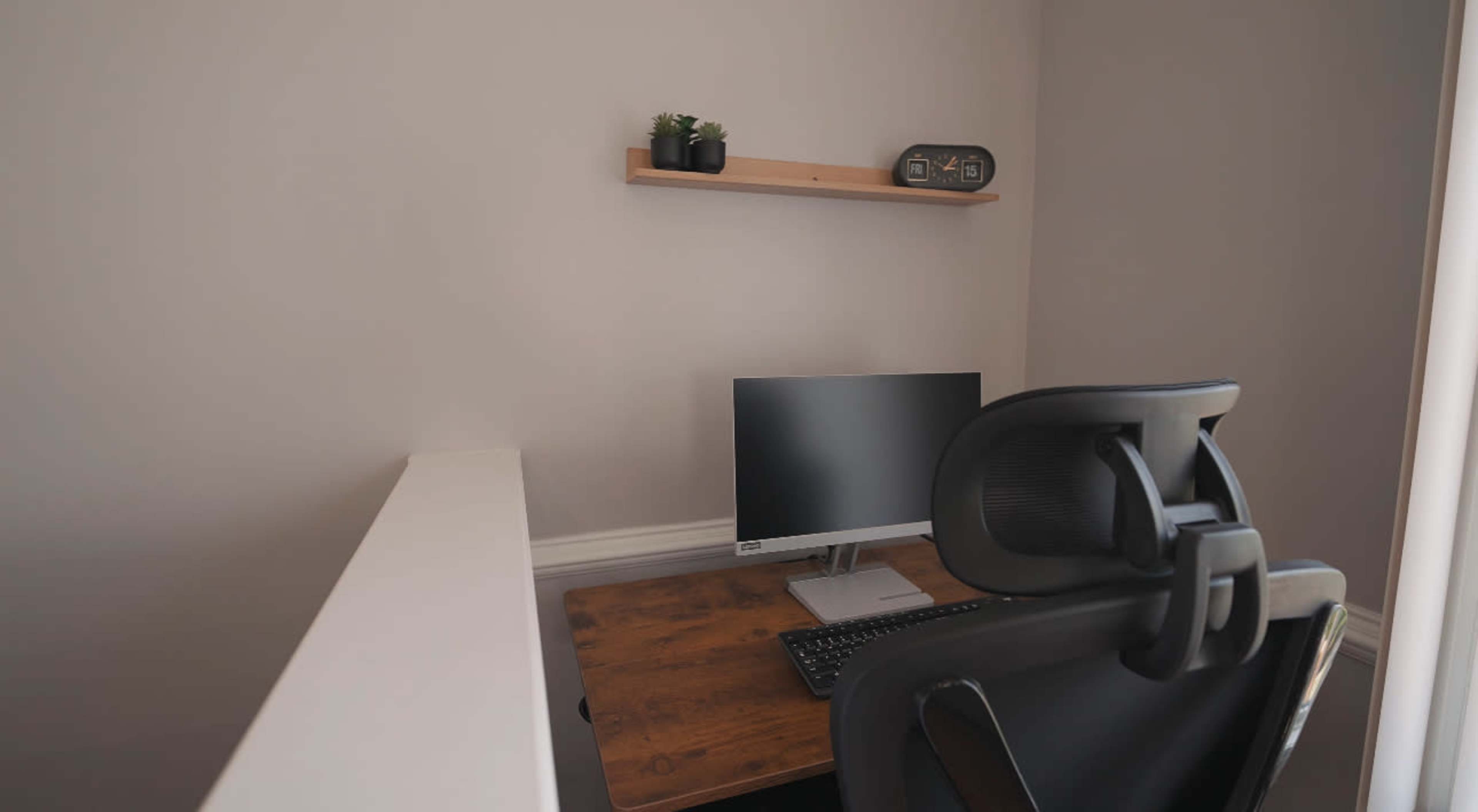 The image shows a minimalist workspace featuring a computer monitor, a keyboard, an office chair, and a shelf with a small plant.