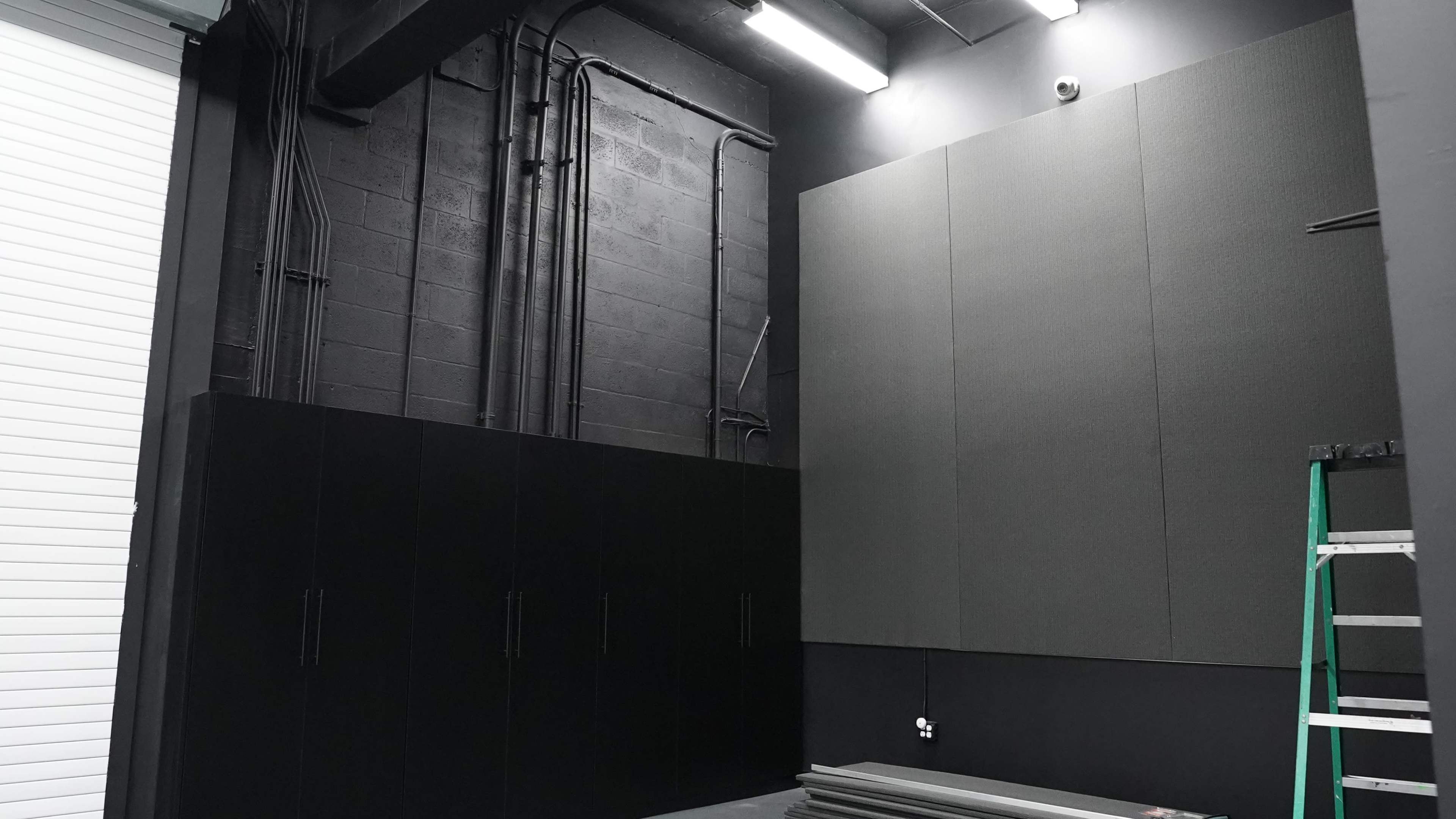 The image shows a dimly lit room with black walls, cabinets, and acoustic panels on the upper section, alongside visible electrical wiring and a ladder.