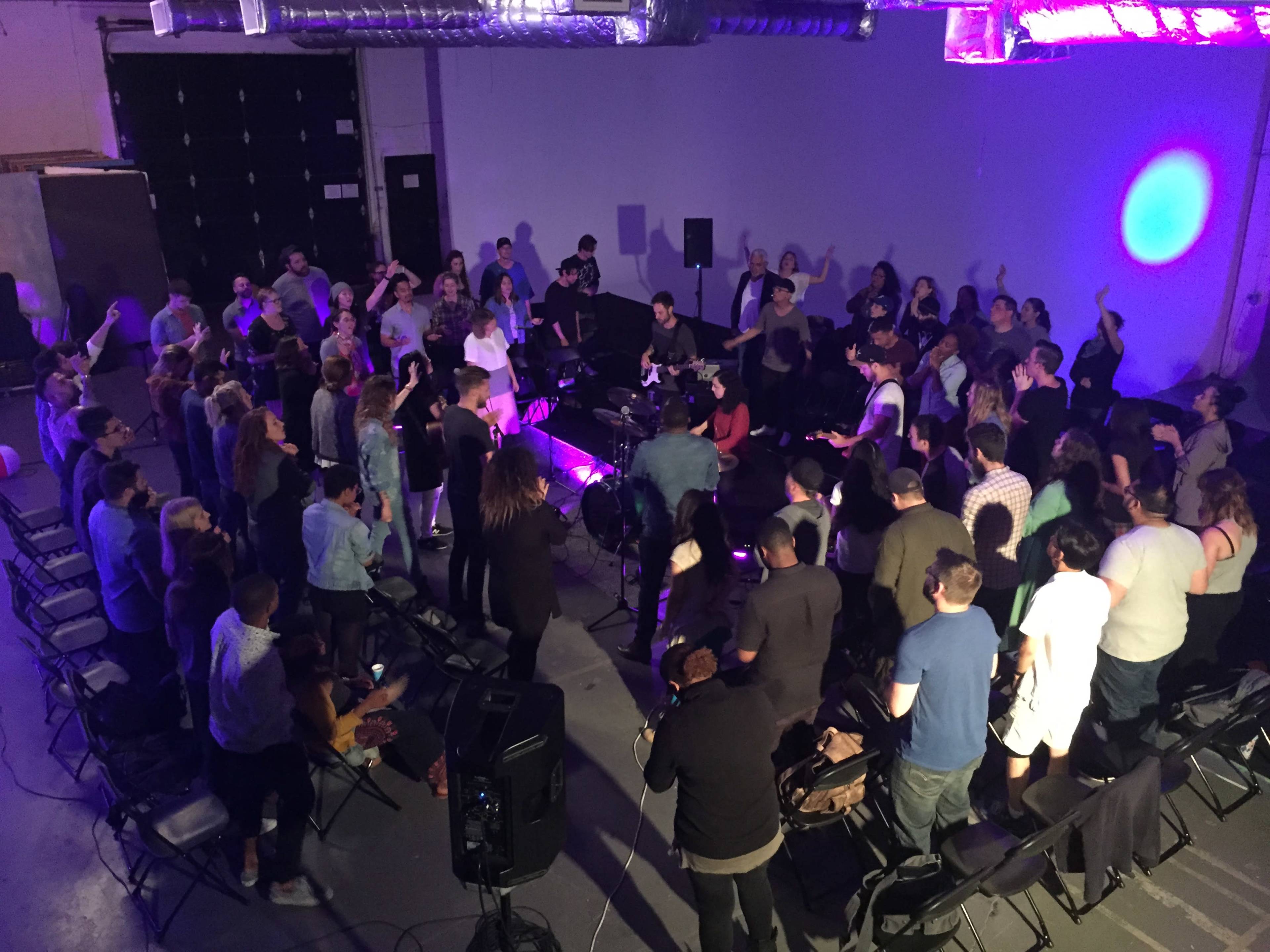 A group of people stands in a dimly lit space, gathered around musicians performing on a small stage.