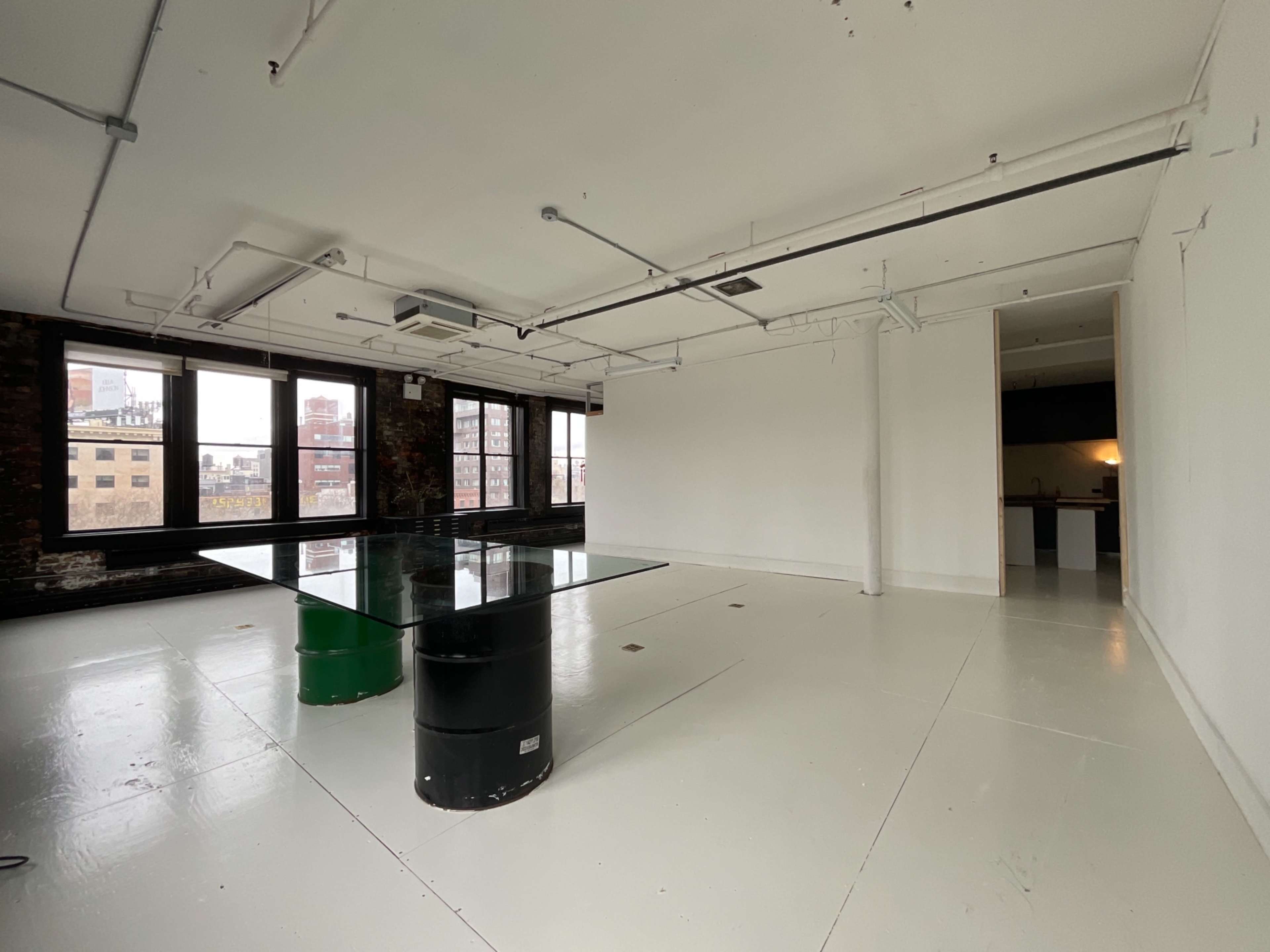 The image shows an empty room with concrete walls, a glass table on top of a black barrel, and large windows overlooking a cityscape.