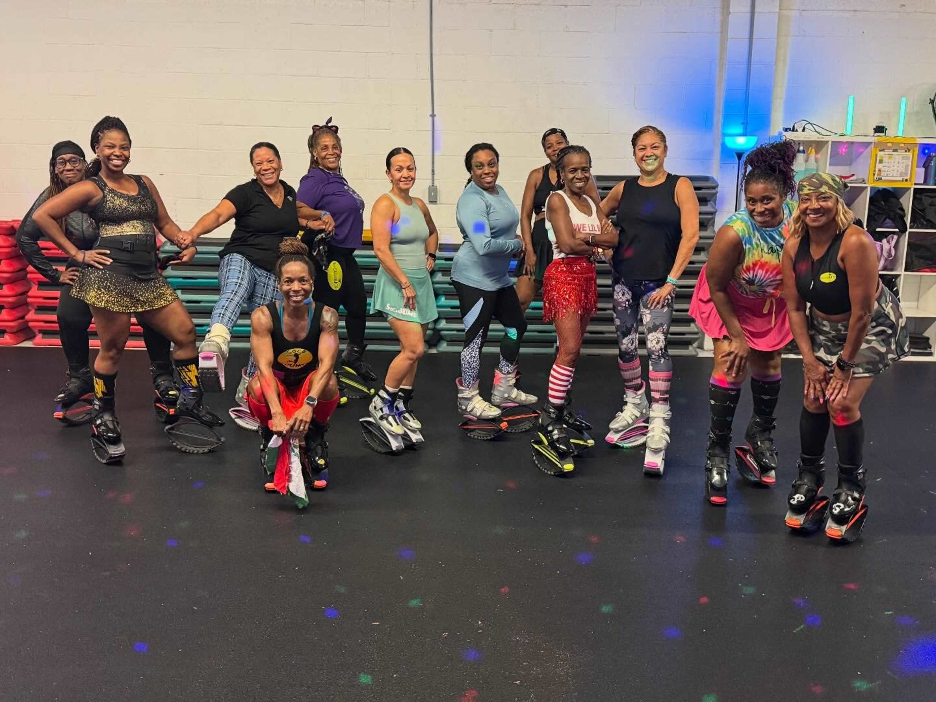 A group of people in colorful workout attire poses with smile while wearing Kangoo Jumps and standing on a gym floor with exercise equipment in the background.
