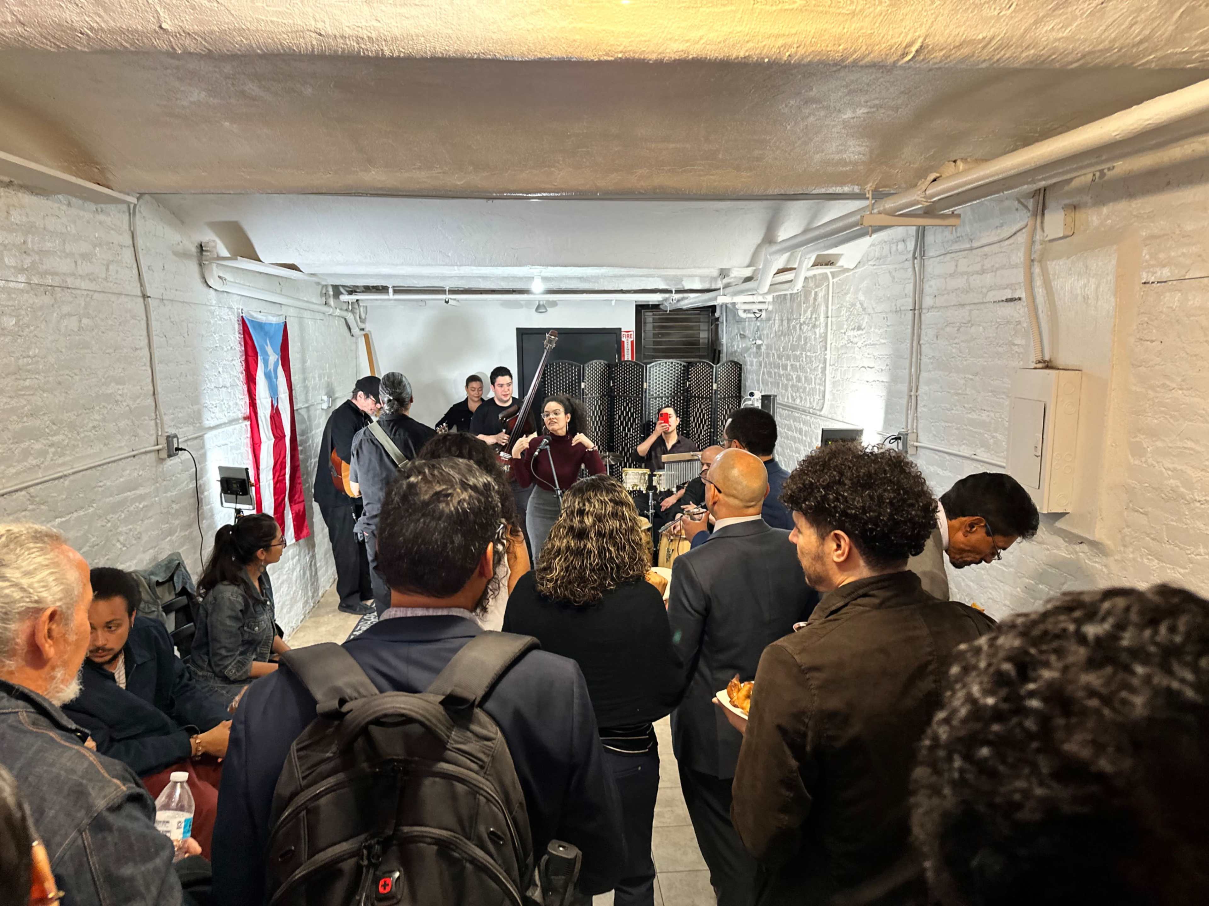 A group of people is gathered in a basement setting, some seated and others standing, as a woman speaks at a microphone in front of a small stage.