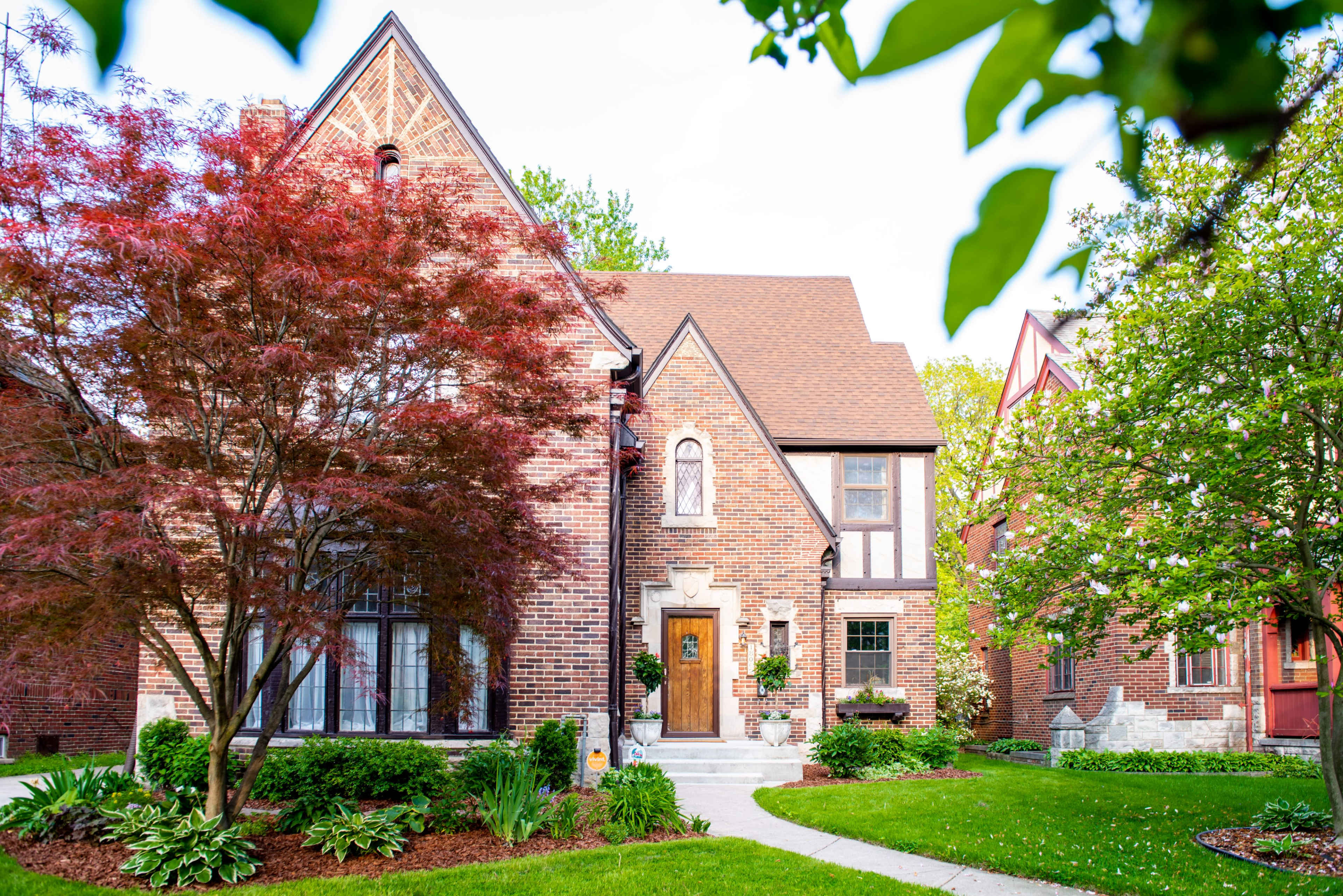 A brick house with a steeply pitched roof is surrounded by green landscaping and a small red maple tree.