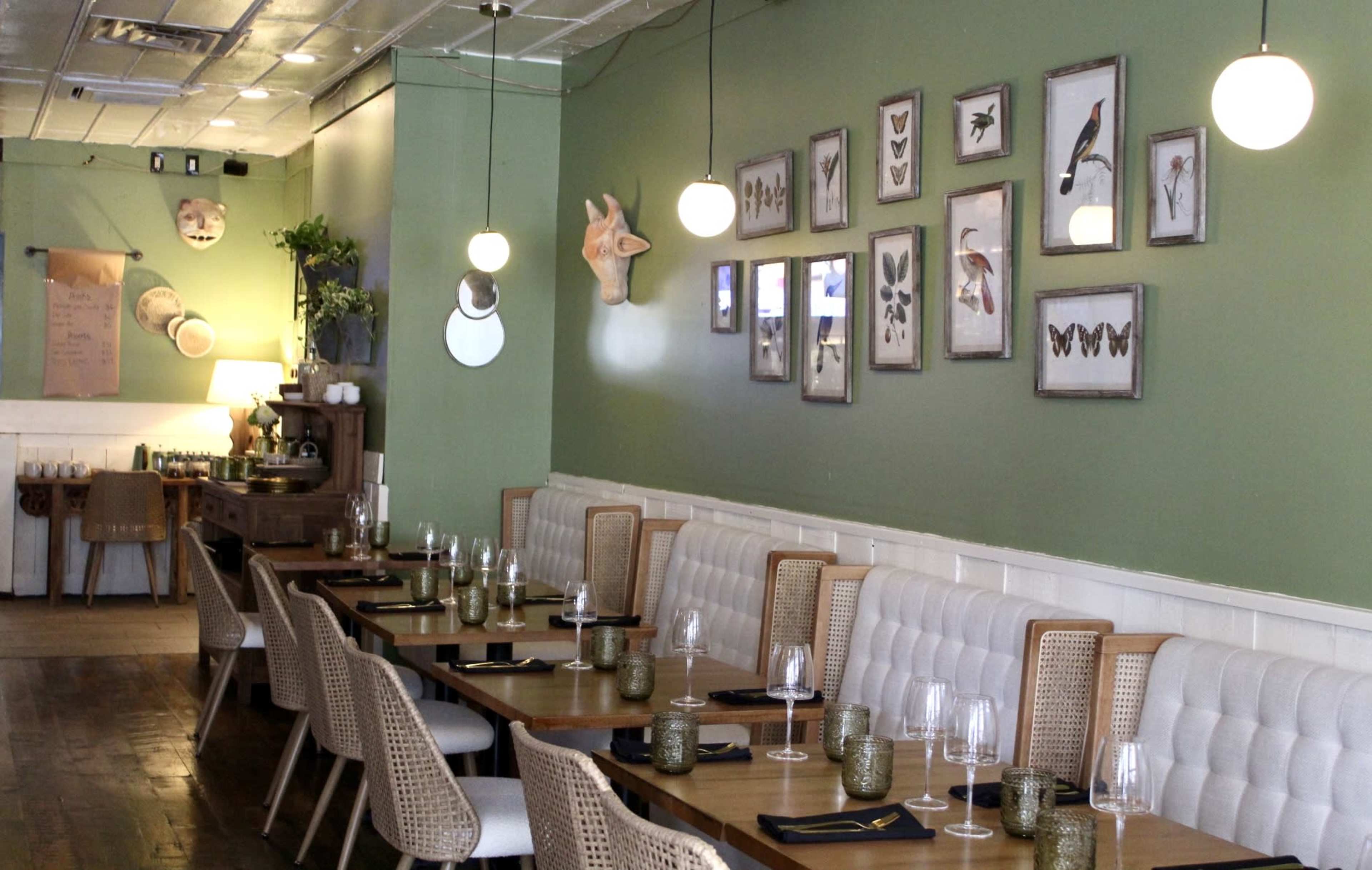 The image shows a restaurant interior featuring wooden tables set with glassware, a green wall adorned with framed nature illustrations, and unique decorative items.