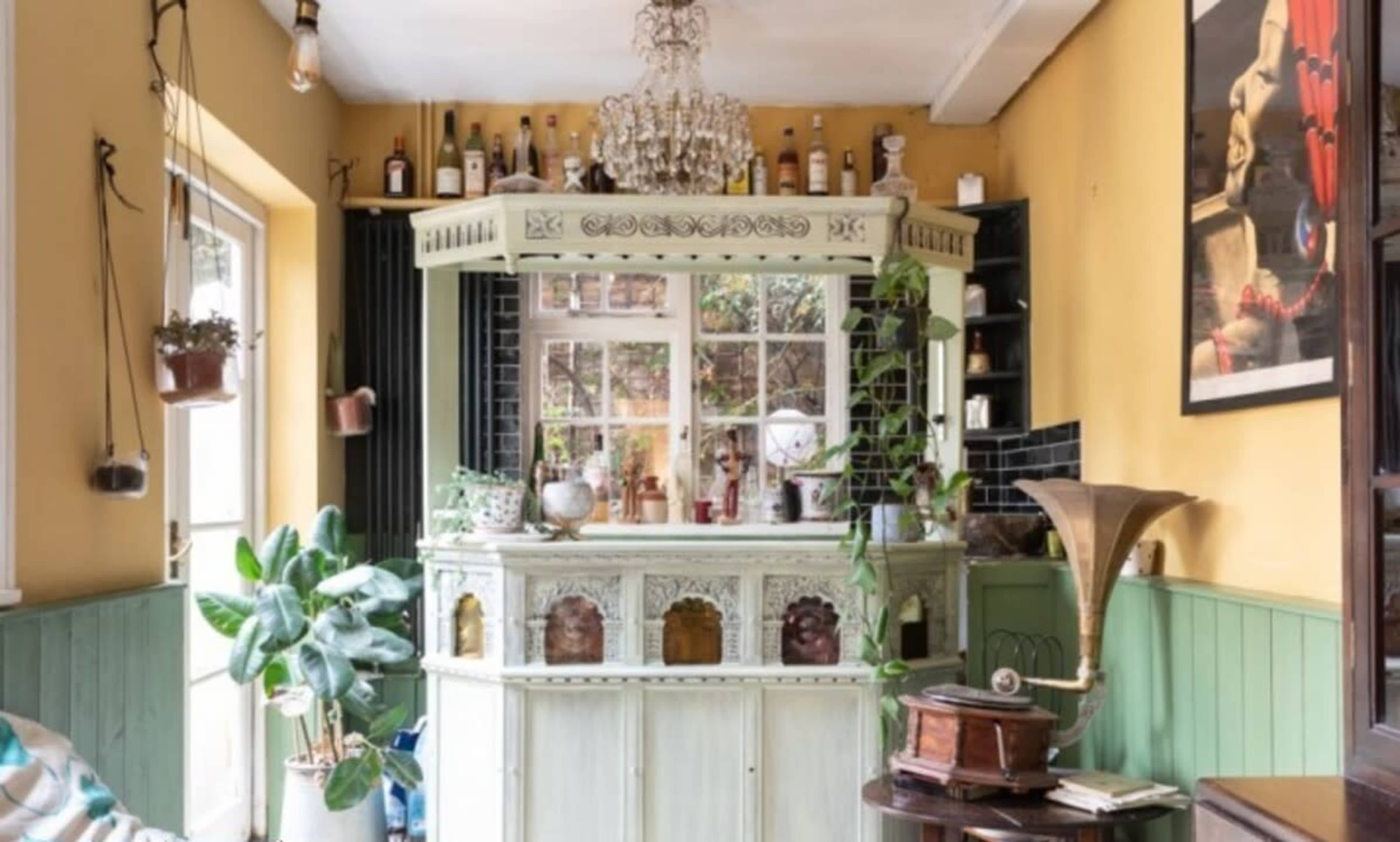The image shows a vintage-style bar area featuring a decorative wooden counter, various bottles on shelves, and a chandelier overhead, surrounded by plants and artwork.