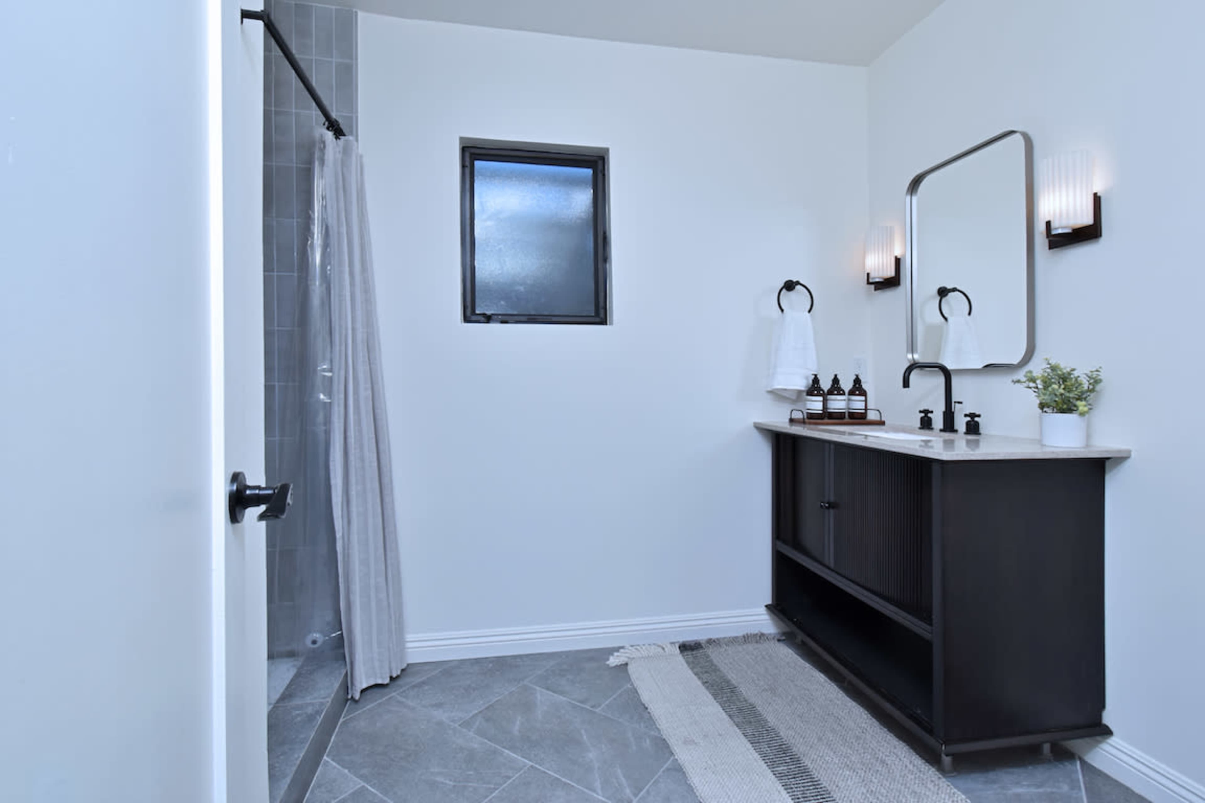 The image shows a modern bathroom featuring a vanity with a sink, a shower area with a curtain, and a window.