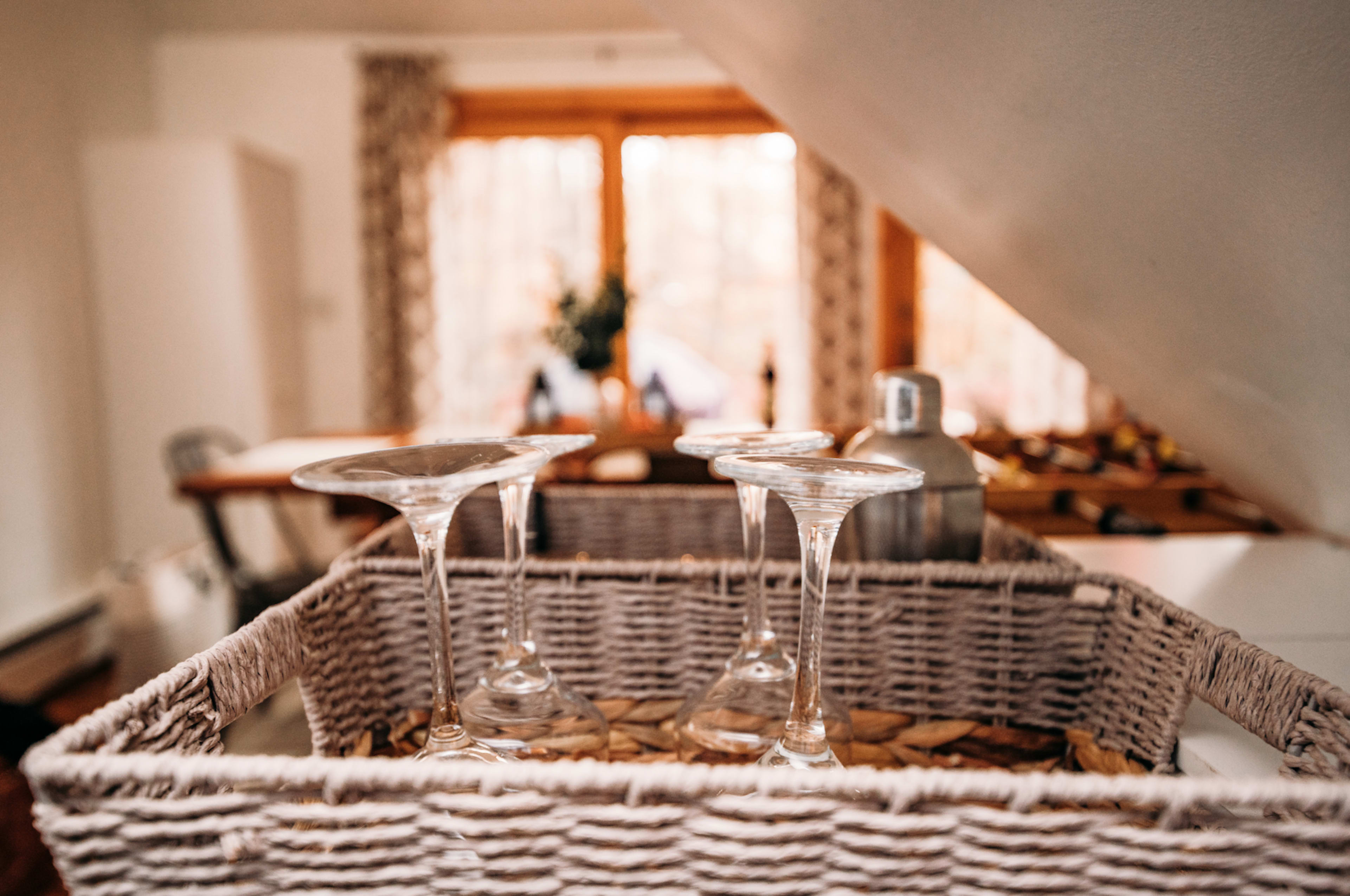 A basket containing three glass cocktail glasses is placed on a surface in a warmly lit, cozy interior space.