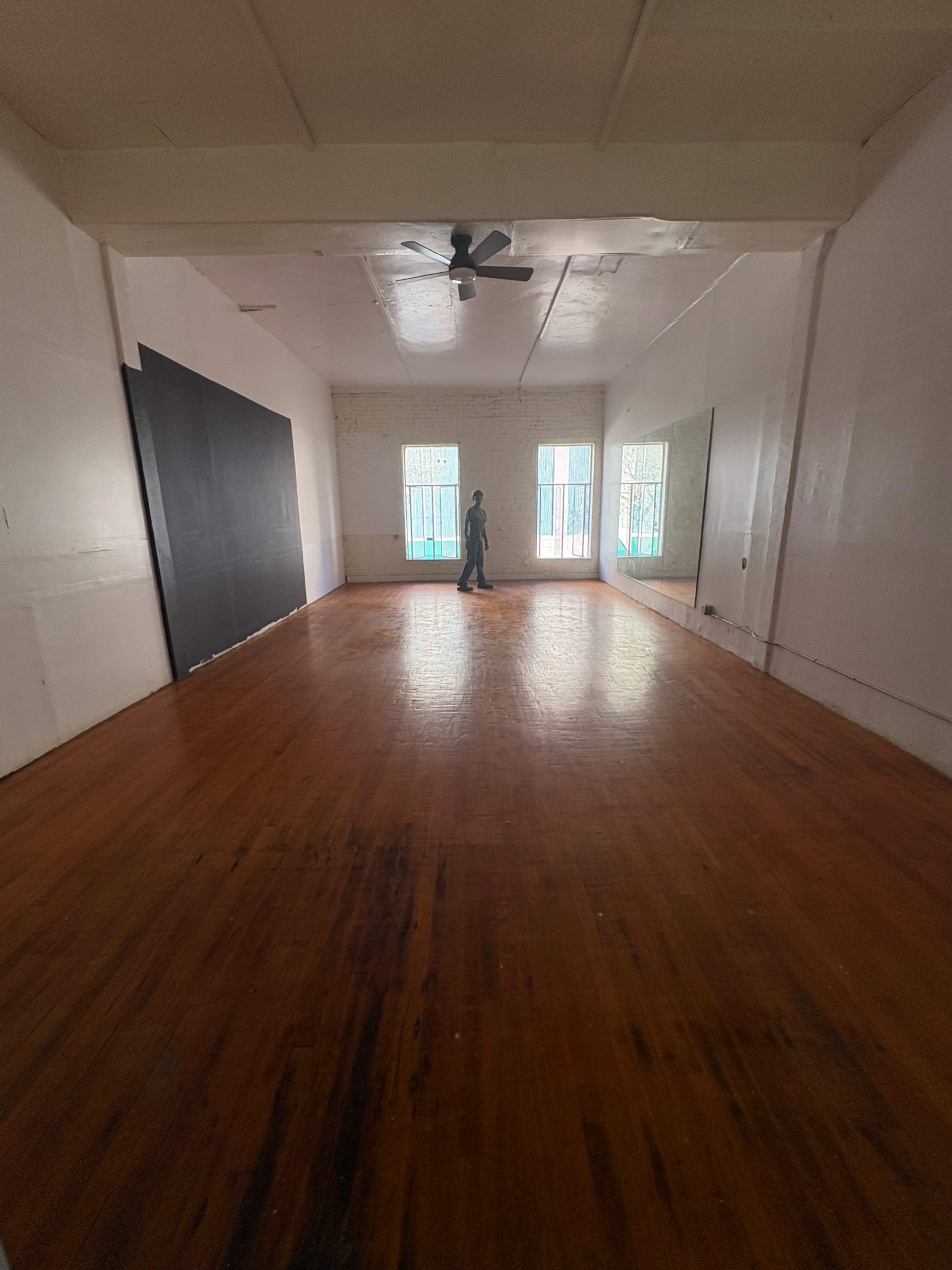 A person stands alone in a spacious, empty room with wooden floors and two large windows casting light.