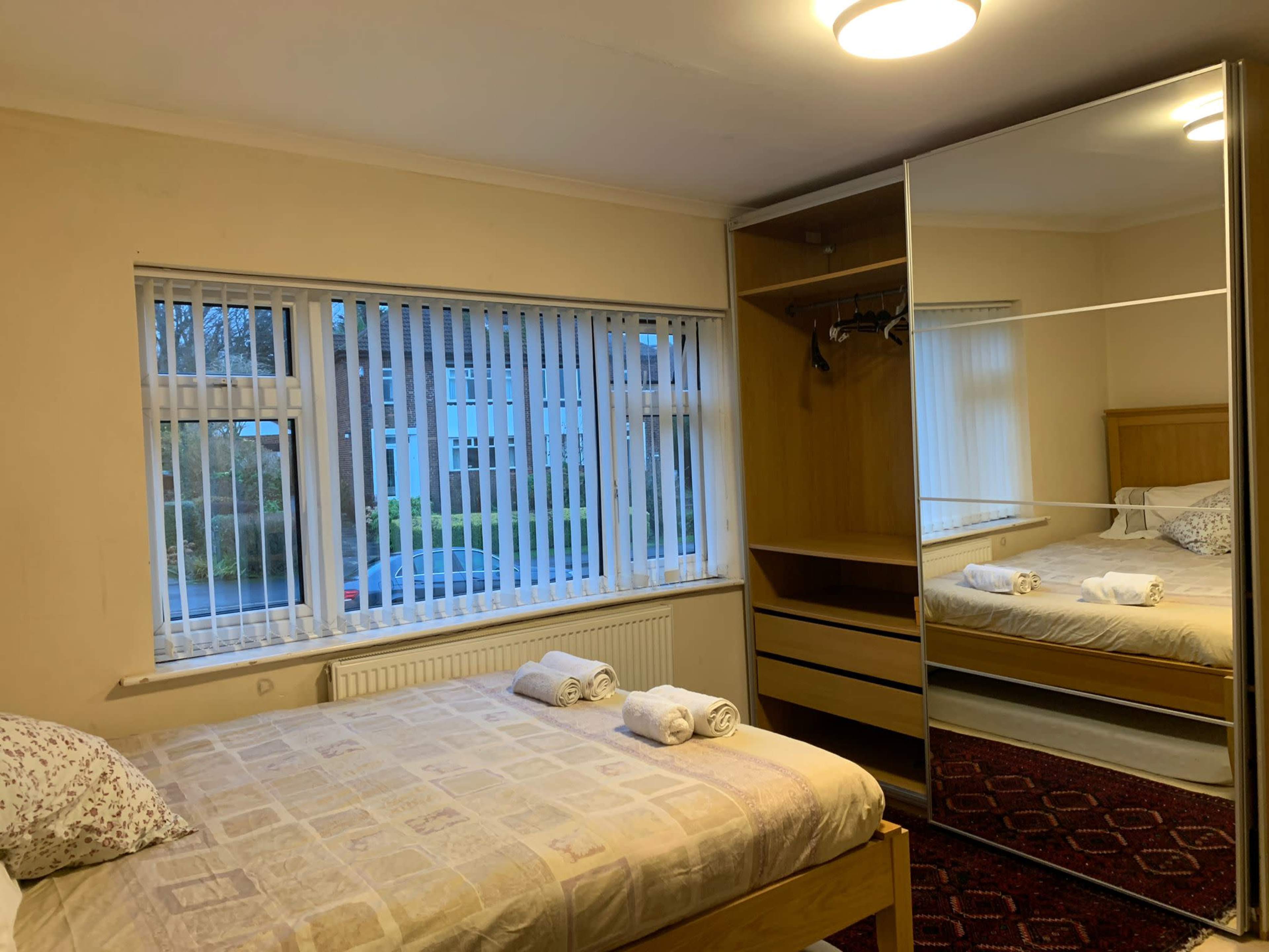 A bedroom features a bed with neatly placed towels, a wardrobe with mirrored doors, and a large window with vertical blinds allowing natural light in.