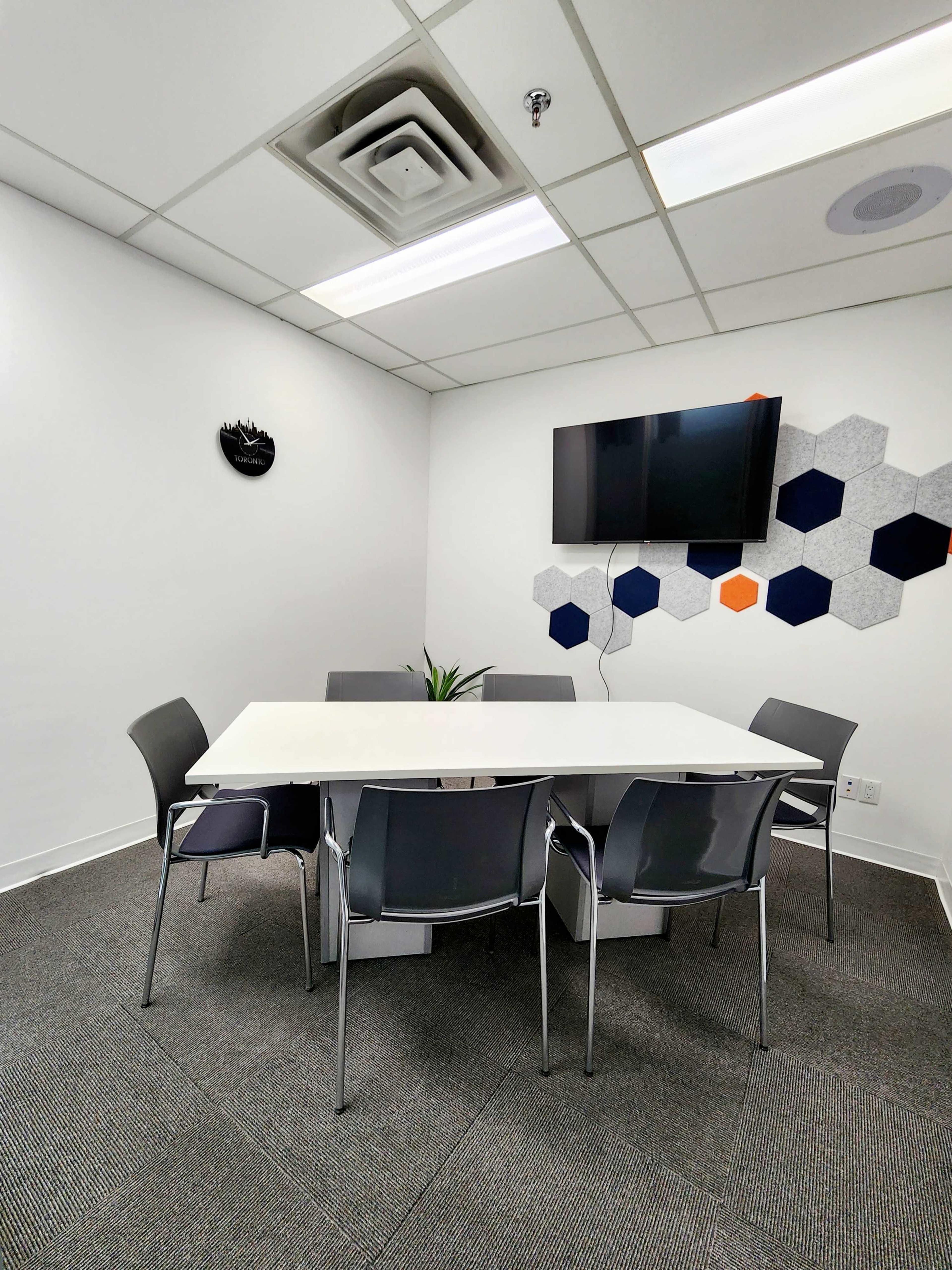 A conference room features a white table surrounded by six gray chairs, a wall-mounted TV, and hexagonal wall decor.