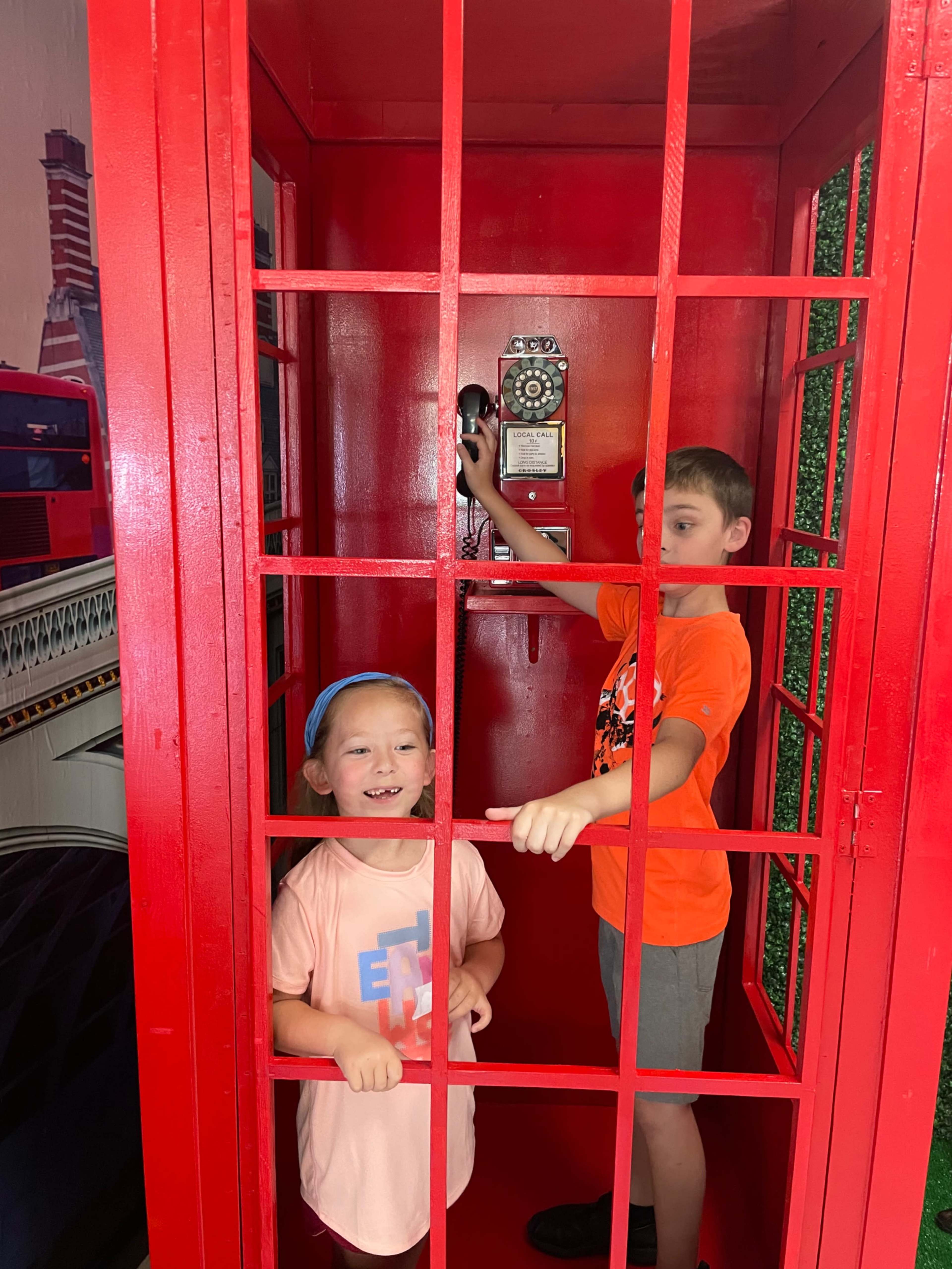 Two children are inside a red telephone booth, with one holding a phone and the other smiling at the camera.