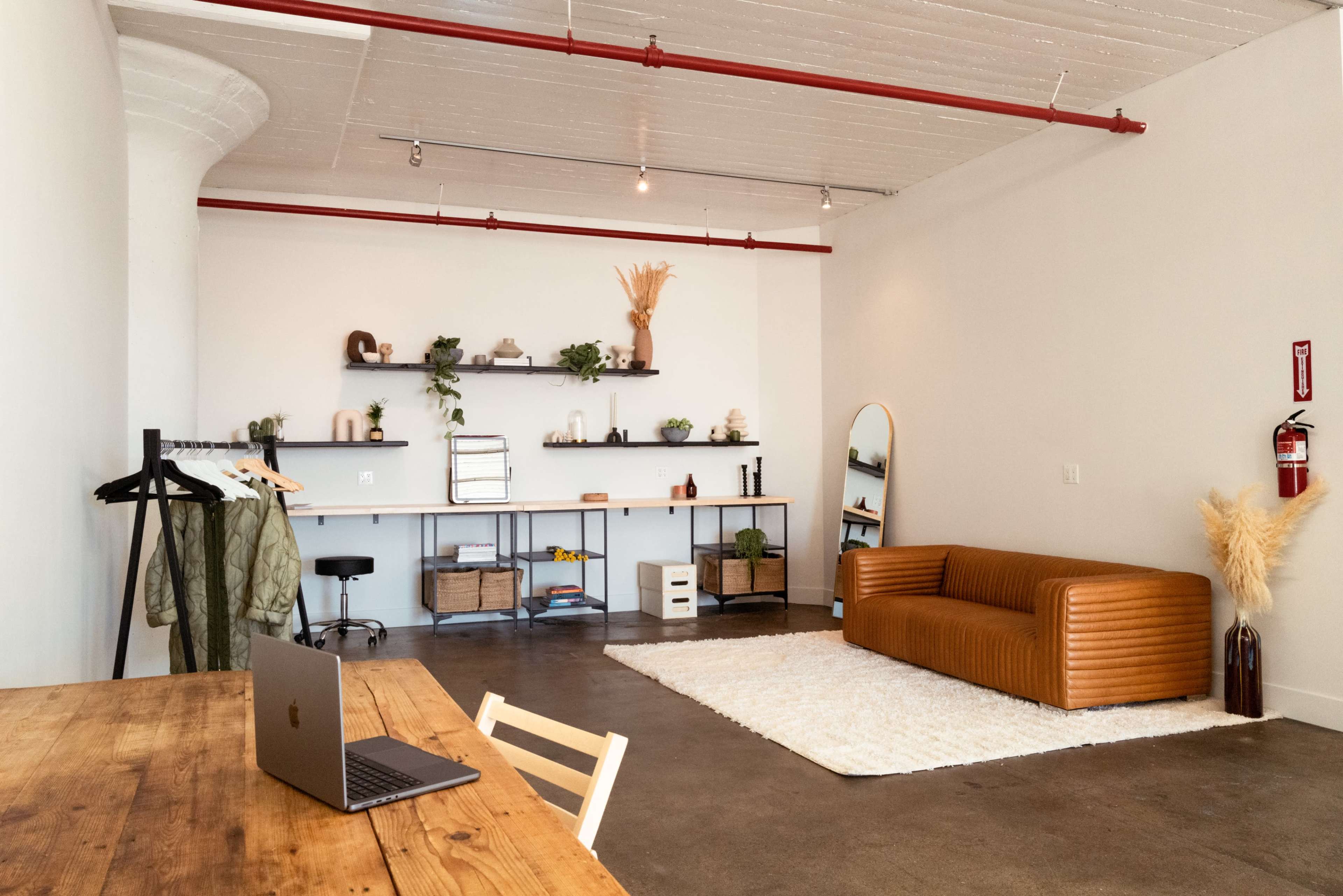 A modern, minimalistic interior with a brown leather couch, a wooden table with a laptop, and a shelving unit decorated with plants and various items.