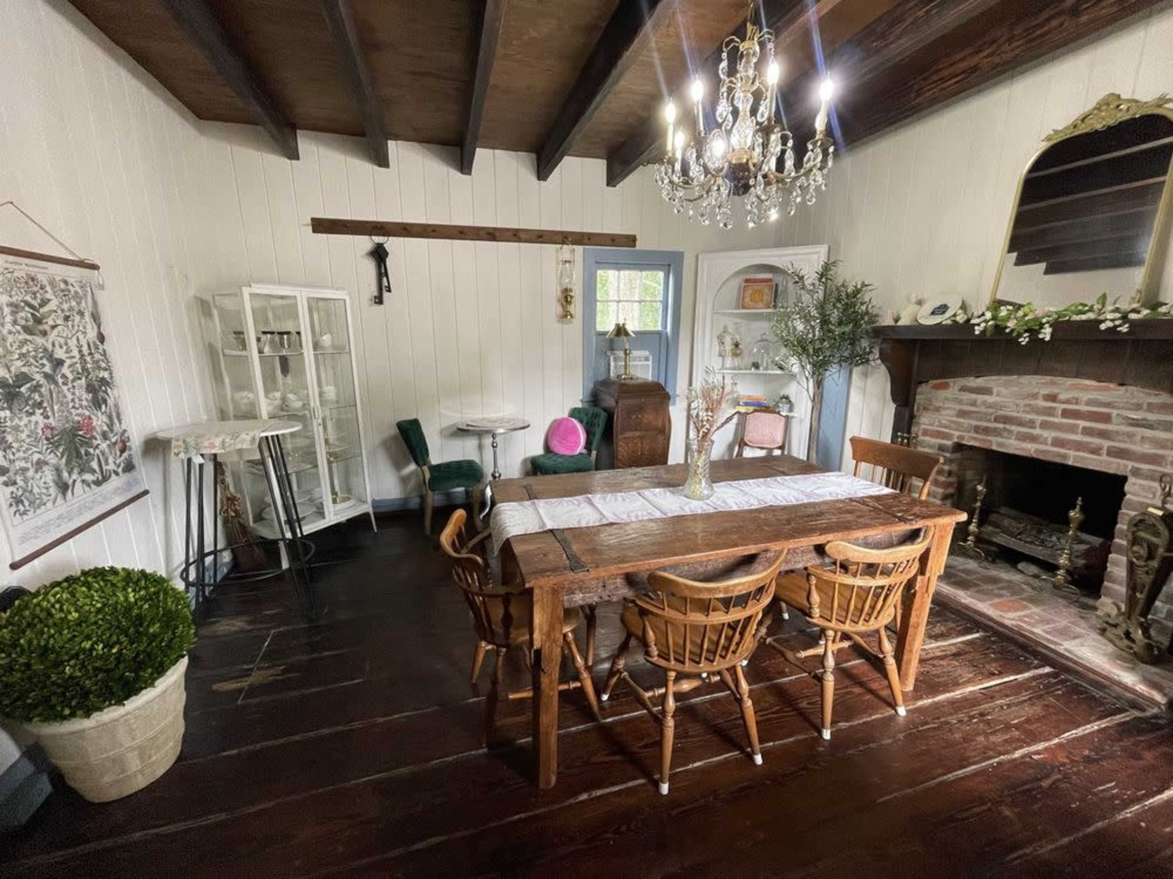 The image shows a dining room with a wooden table surrounded by wicker chairs, a brick fireplace, and various decorative elements including a chandelier and houseplants.